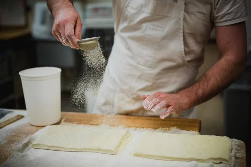 Person sprinkling flour onto dough pieces on a wooden surface in a kitchen.