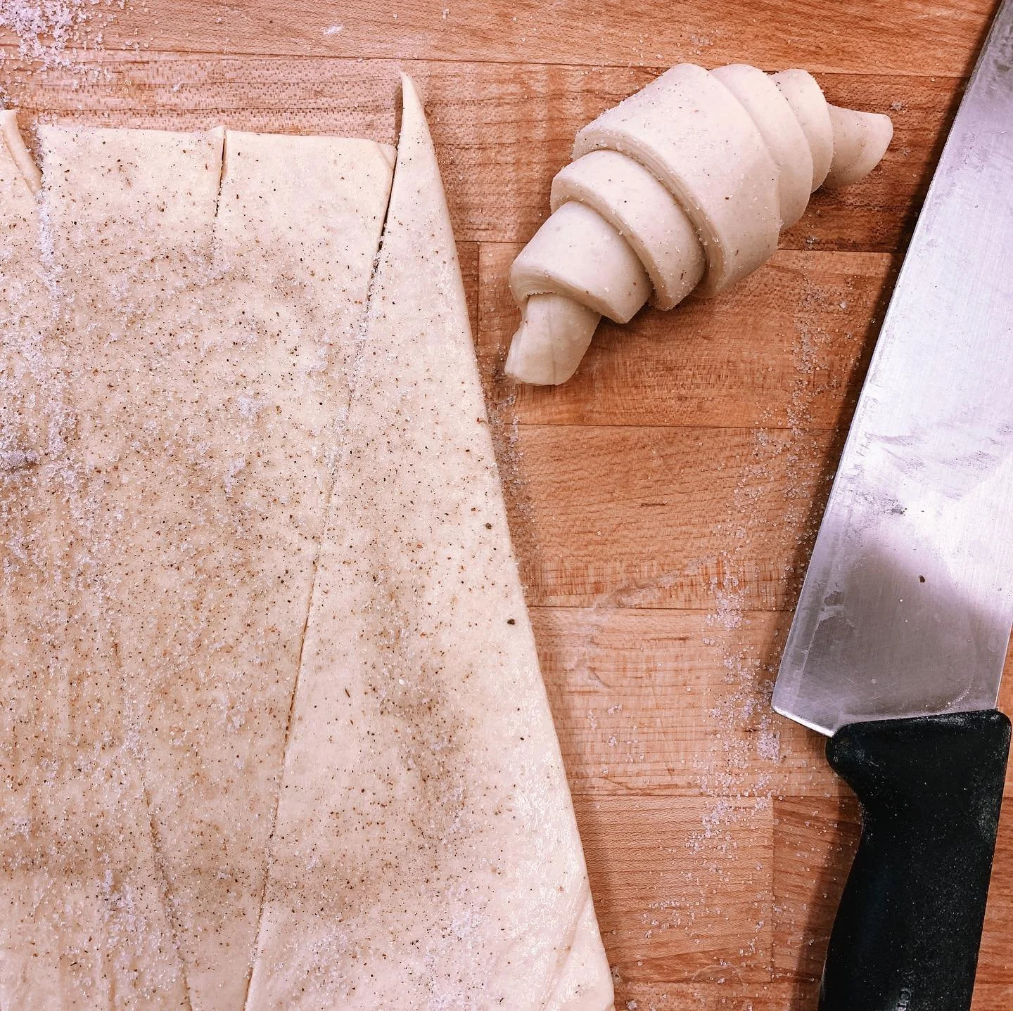 A wooden cutting board with a partially rolled out piece of dough and a large kitchen knife.