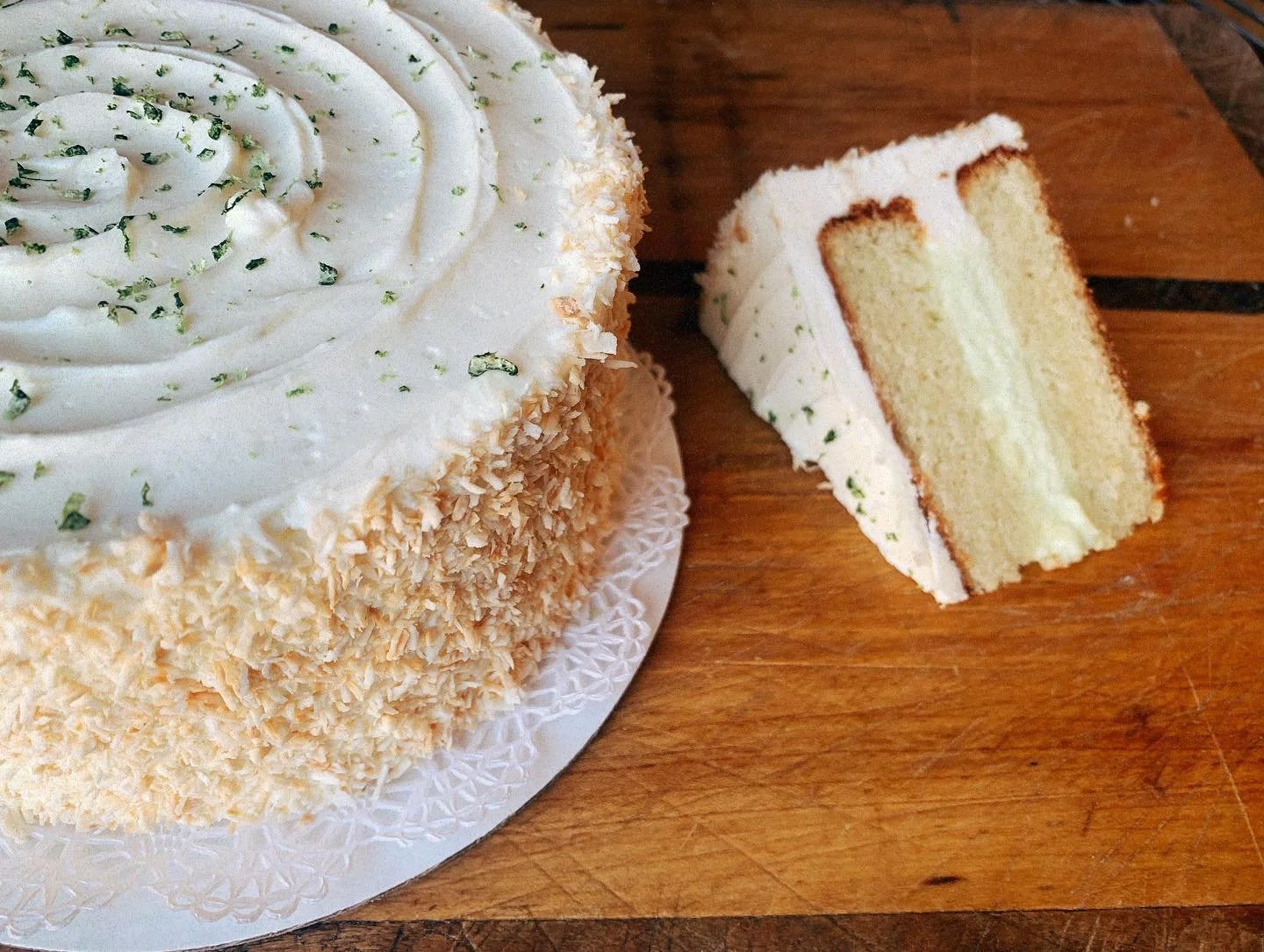 A round cake with white frosting, chopped nuts on the sides, and green sprinkles on top, placed on a white doily on a wooden surface. A slice removed from the cake reveals layers of sponge and cream filling.