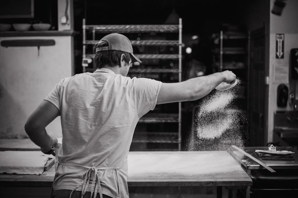 A person wearing a cap and an apron is baking or working with dough in a commercial kitchen. The individual is sifting flour onto a work surface.