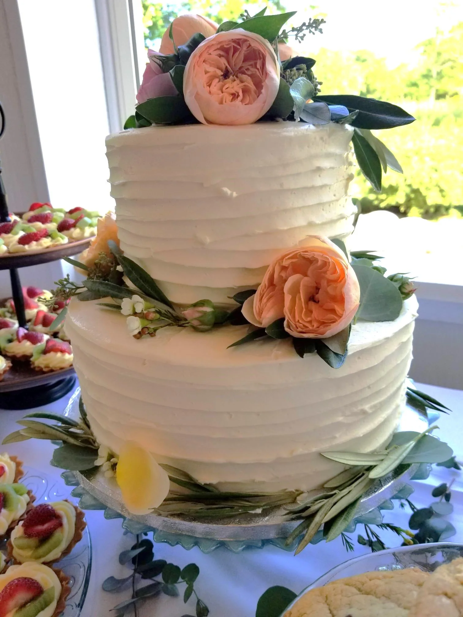 Three-tier white cake decorated with pink and peach roses and green leaves, placed on a silver cake stand. The background shows a window with green trees outside, and there are plates of fruit-topped desserts and cookies on the table.
