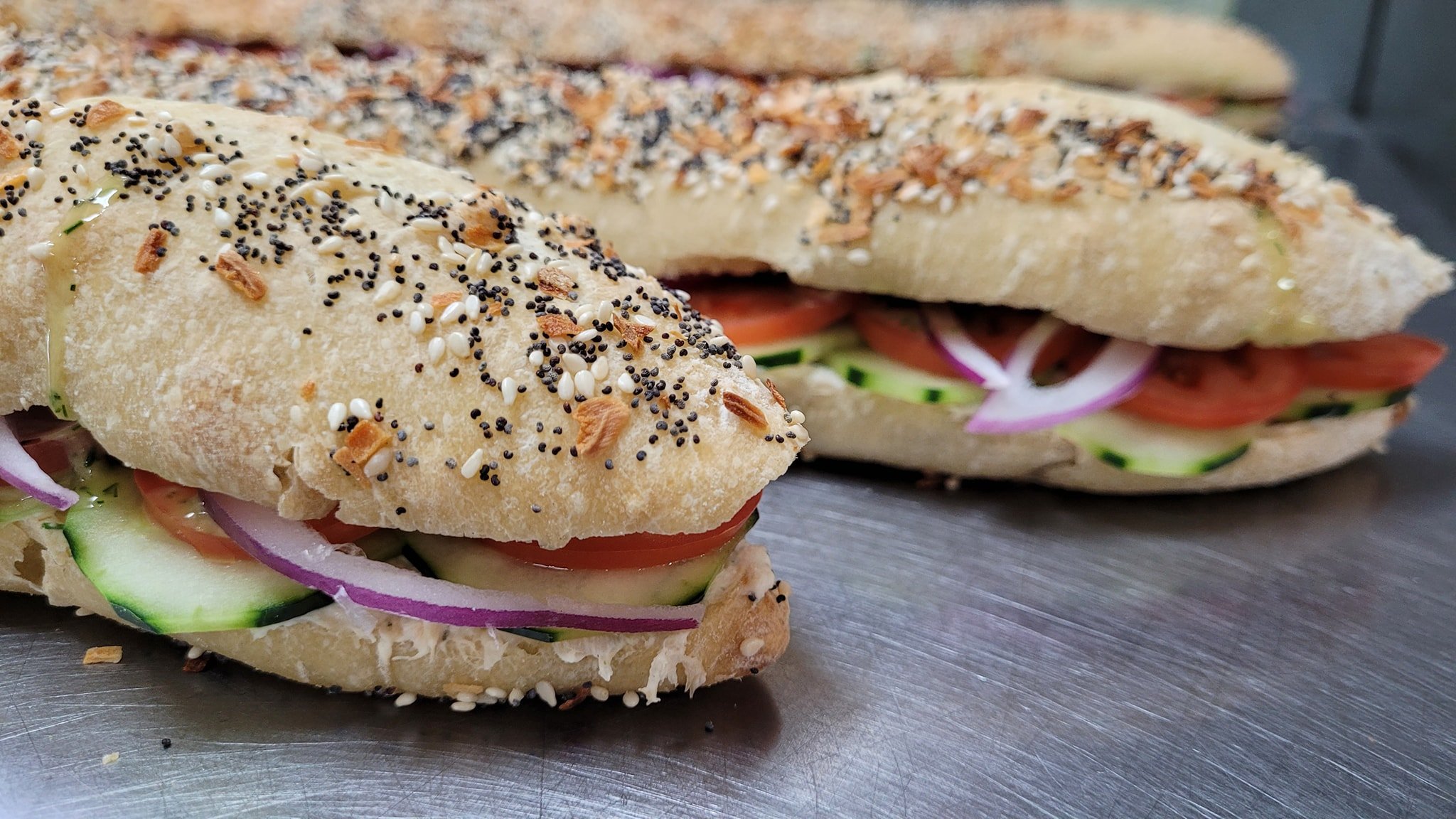 Close-up of a sandwich with sliced cucumbers, tomatoes, red onions, and sprigs of herbs inside a sesame seed bagel.