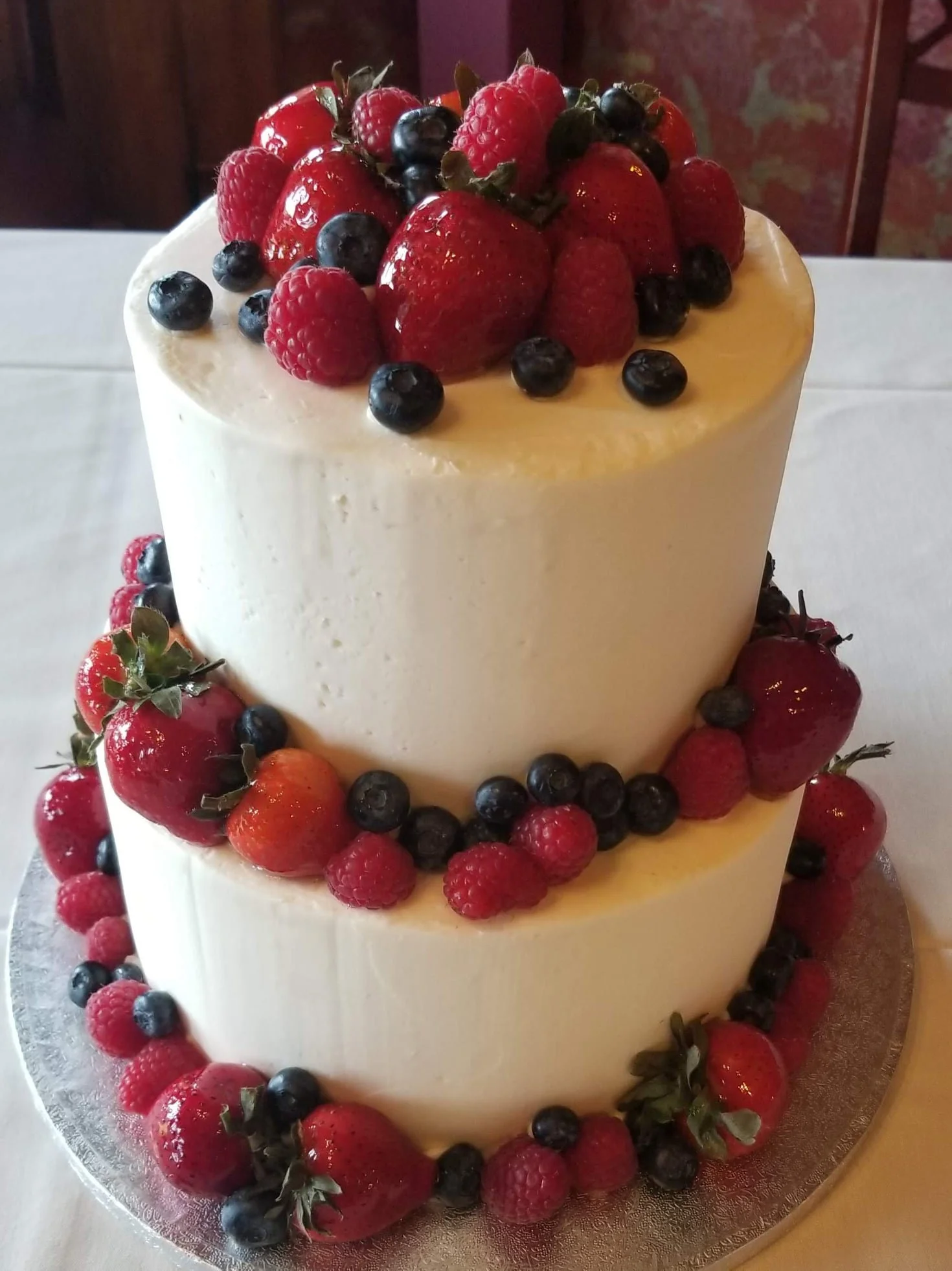 A two-tiered white cake decorated with fresh strawberries, blueberries, and raspberries on top and around the sides.