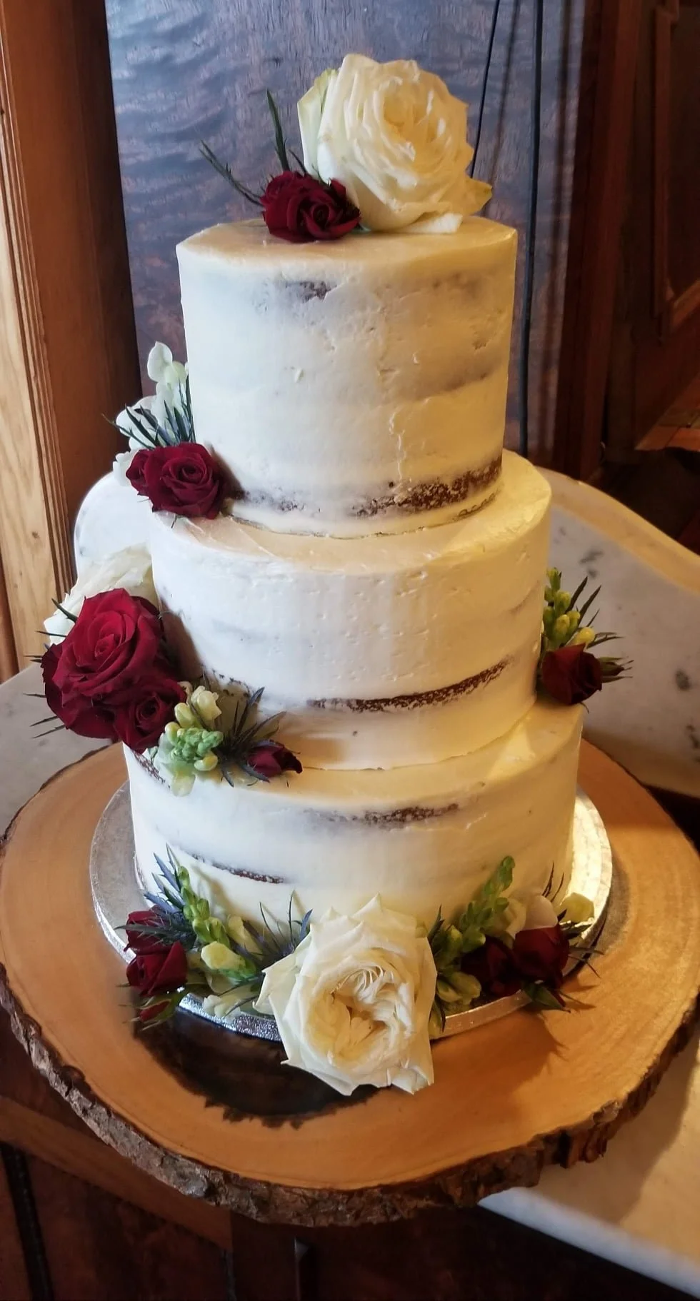 A three-tiered wedding cake with semi-naked frosting, decorated with white and red roses, green buds, and greenery, placed on a wooden slab.