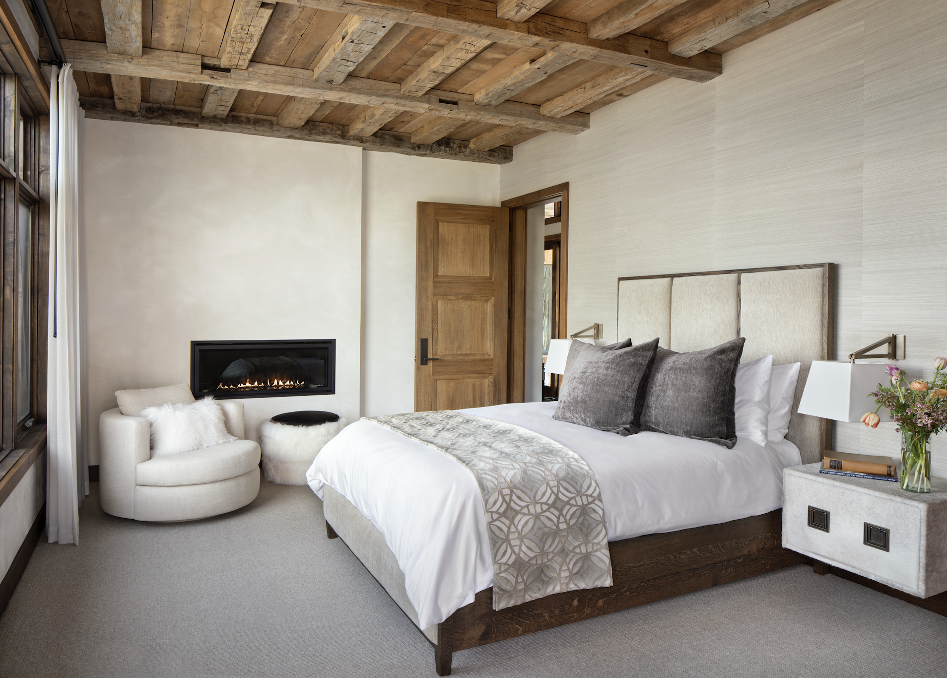 Bedroom with upholstered bed, gas fireplace, reclaimed timber ceiling, and sitting area.