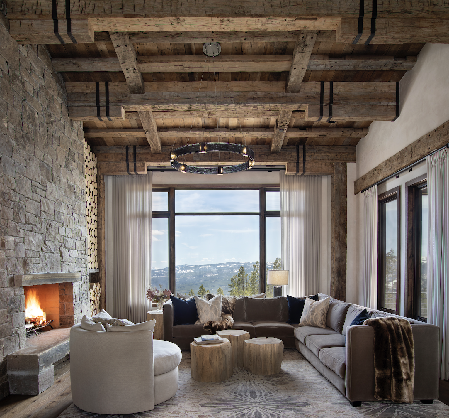 Living room with stone fireplace, reclaimed timber beams, and large windows overlooking mountain views.