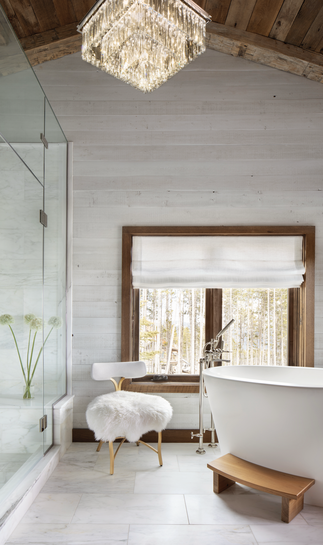 Master bathroom with freestanding soaking tub, marble finishes, and forest views through a wood-framed window.