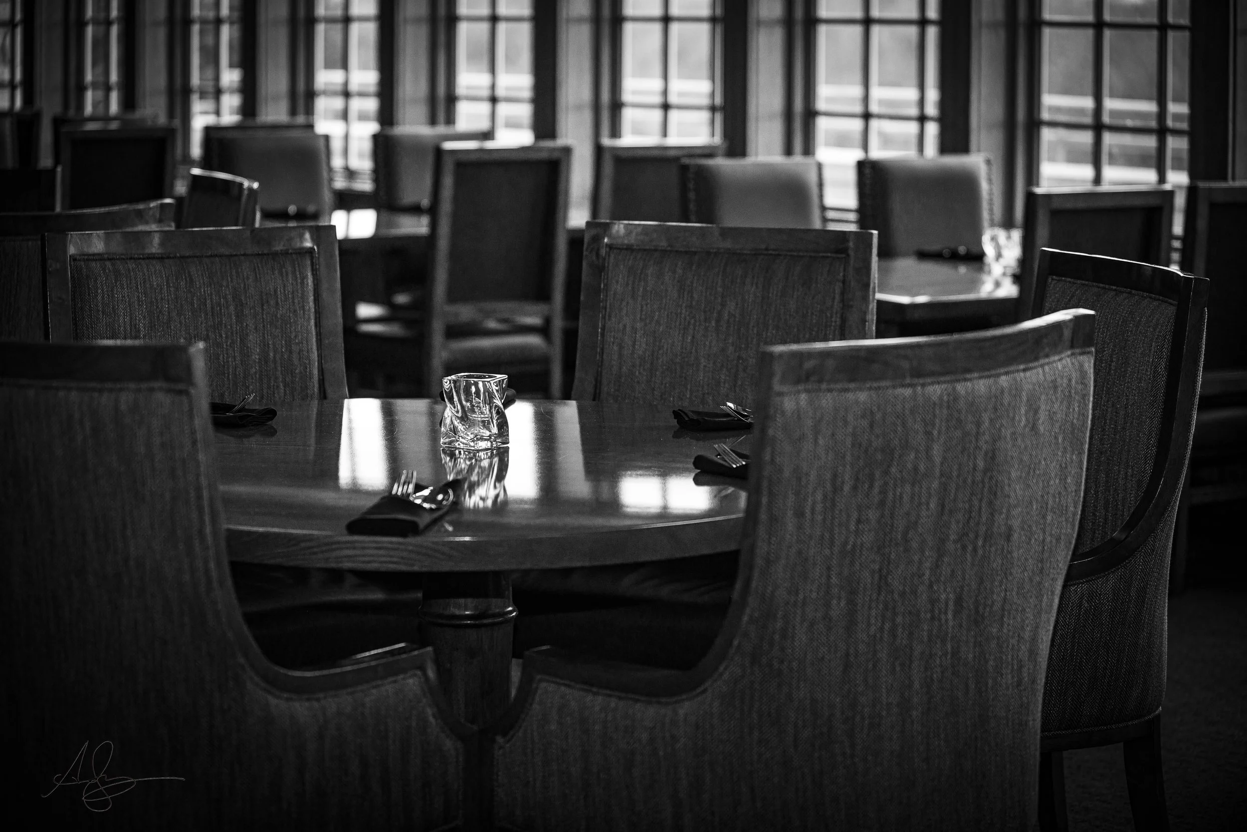 A black and white photograph of a dining area with several empty chairs and tables, set with utensils and glasses, near large windows.