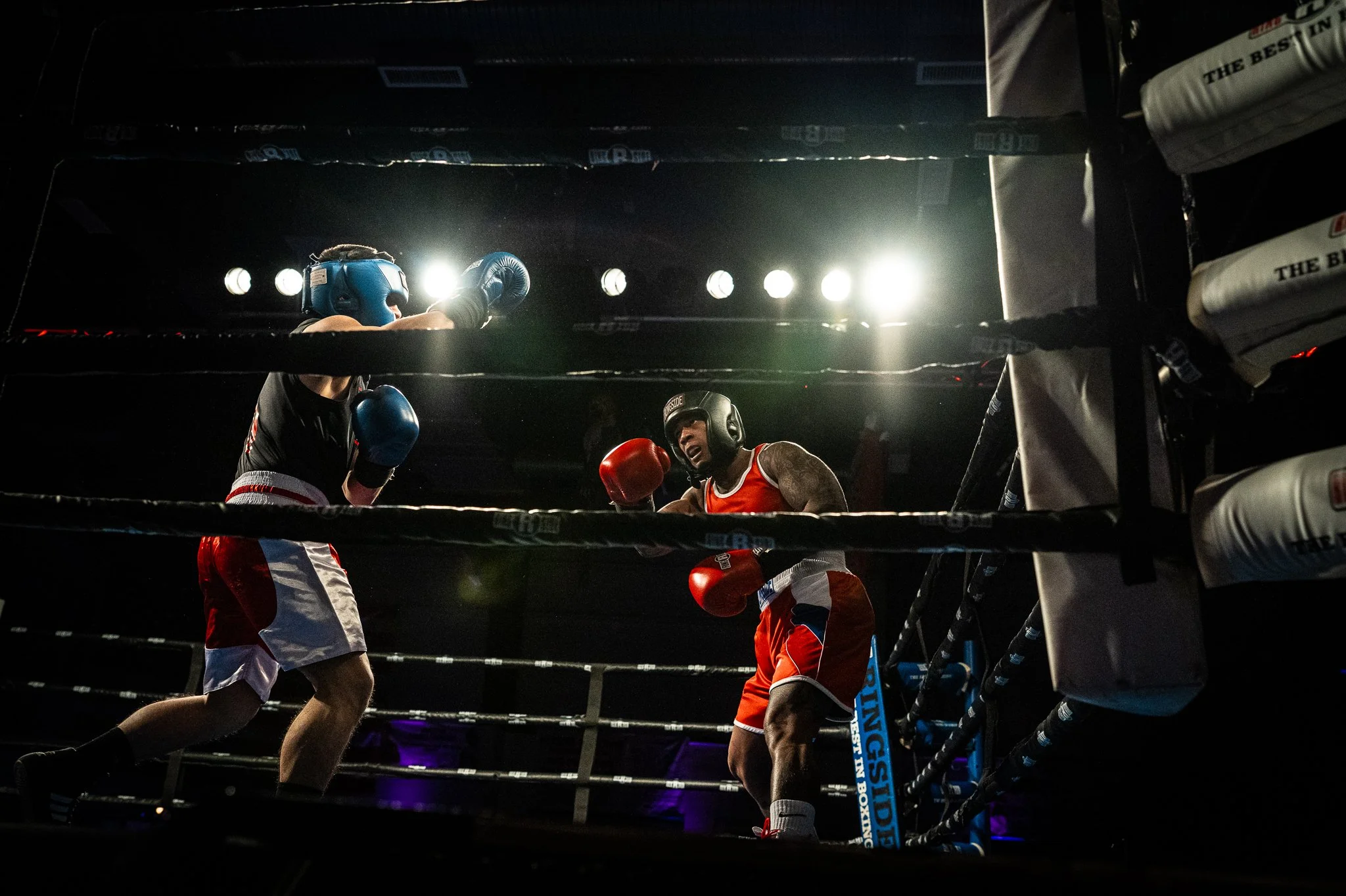 Two male boxers are engaged in a match inside a boxing ring, wearing protective headgear and boxing gloves. One boxer is wearing blue gloves and headgear, while the other is wearing red gloves and headgear. Bright lights are shining from above, illuminating the scene.