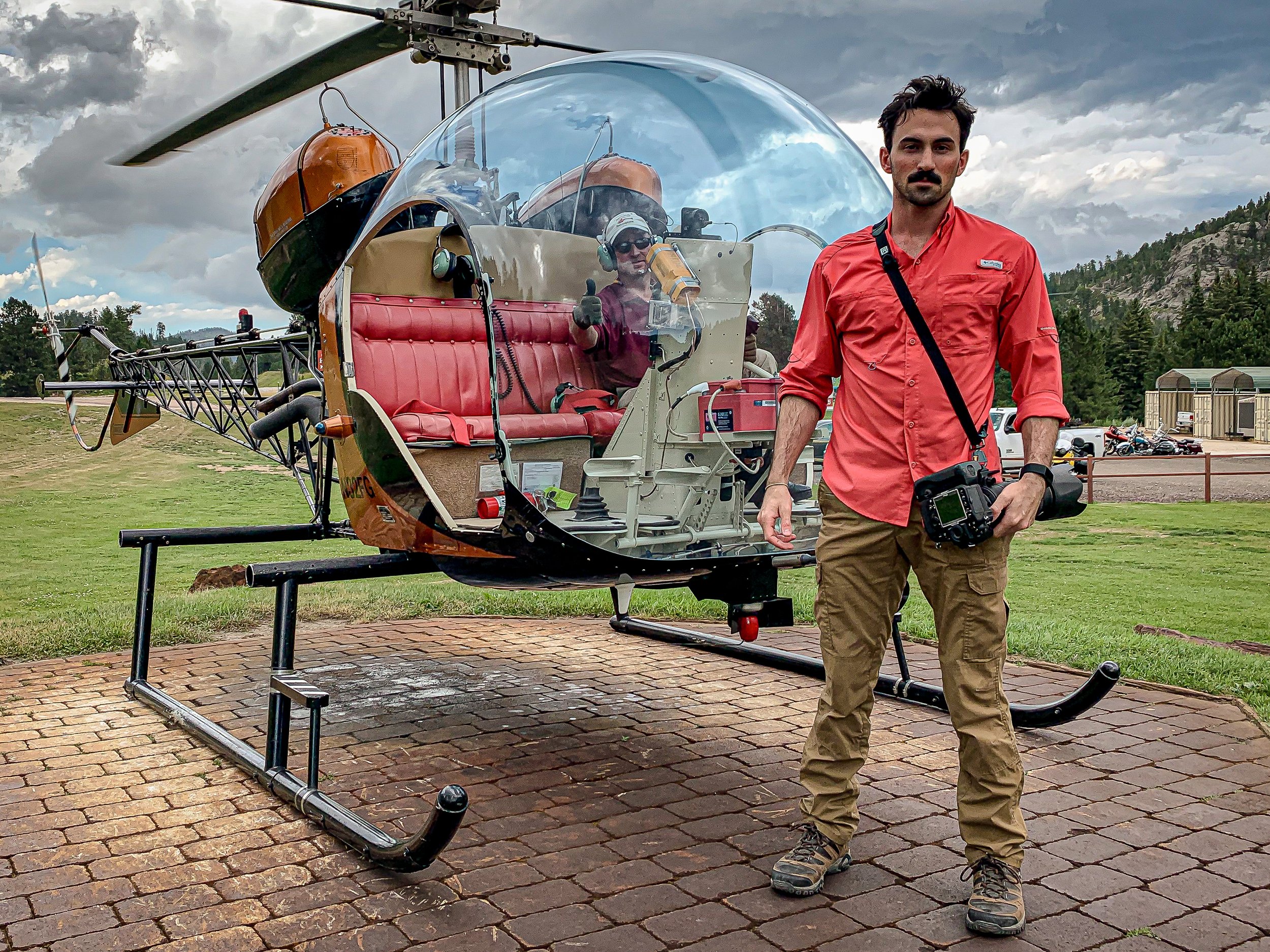 A man in a red shirt and khaki pants standing next to a helicopter on a brick-paved surface outdoors, with cloudy skies and trees in the background.