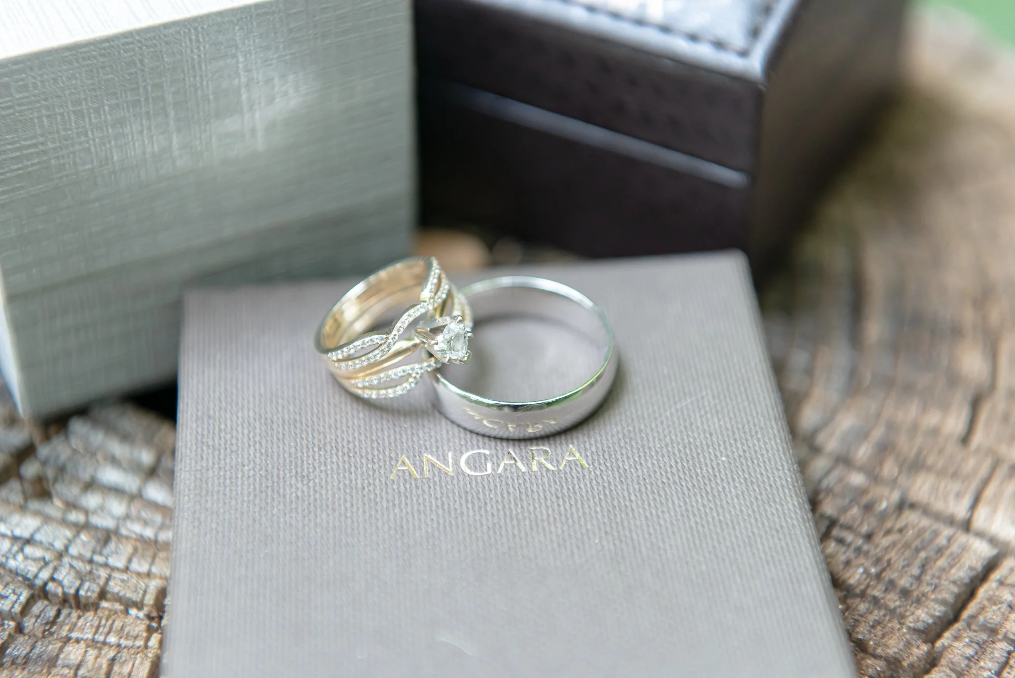 A silver ring with a large central diamond and smaller diamonds along the band, placed on top of a gray ANGARA jewelry box, with another silver ring beside it, on a textured outdoor surface.
