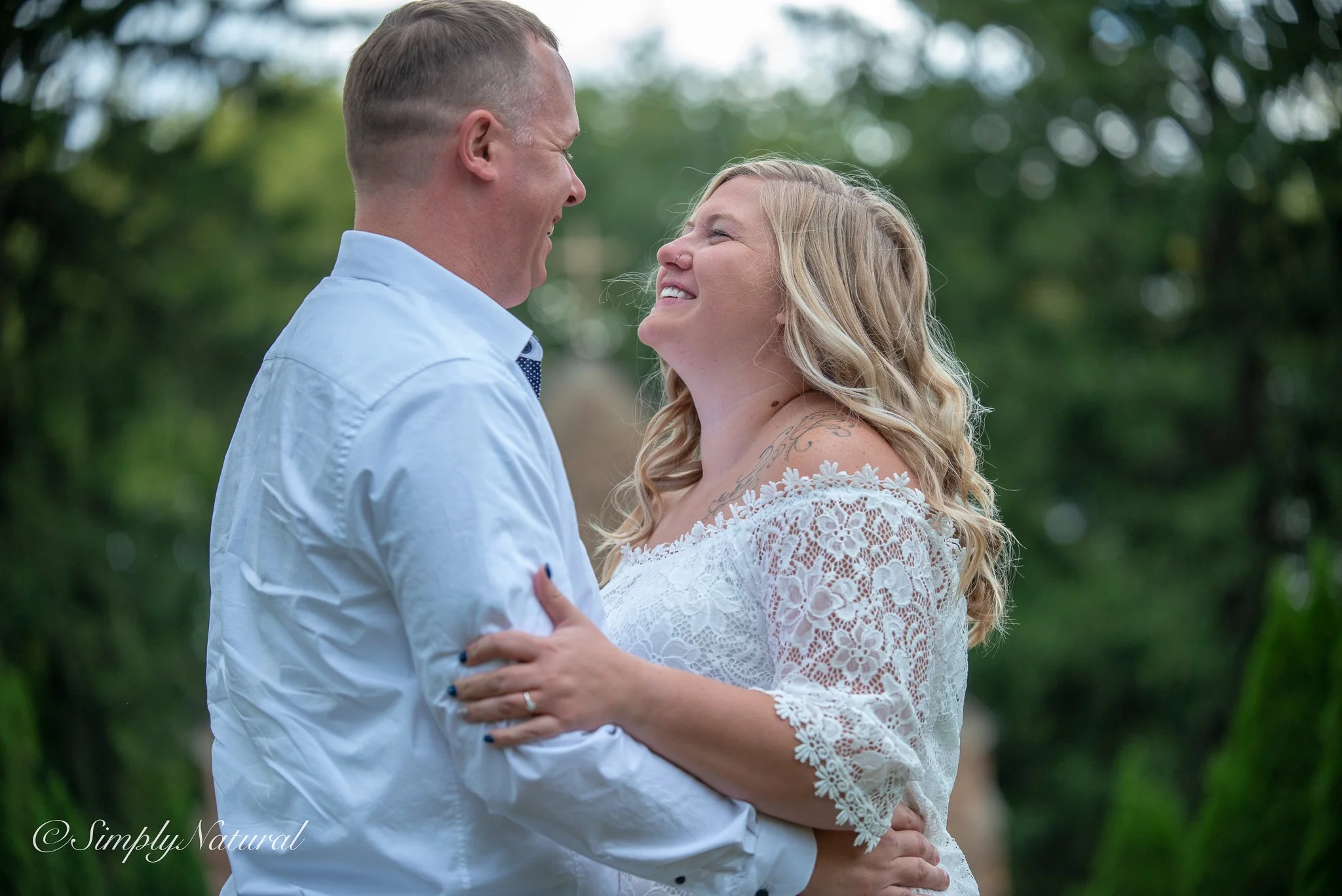 A couple dancing outdoors, smiling, with trees in the background. The woman wears a white lace dress, and the man wears a light blue shirt.