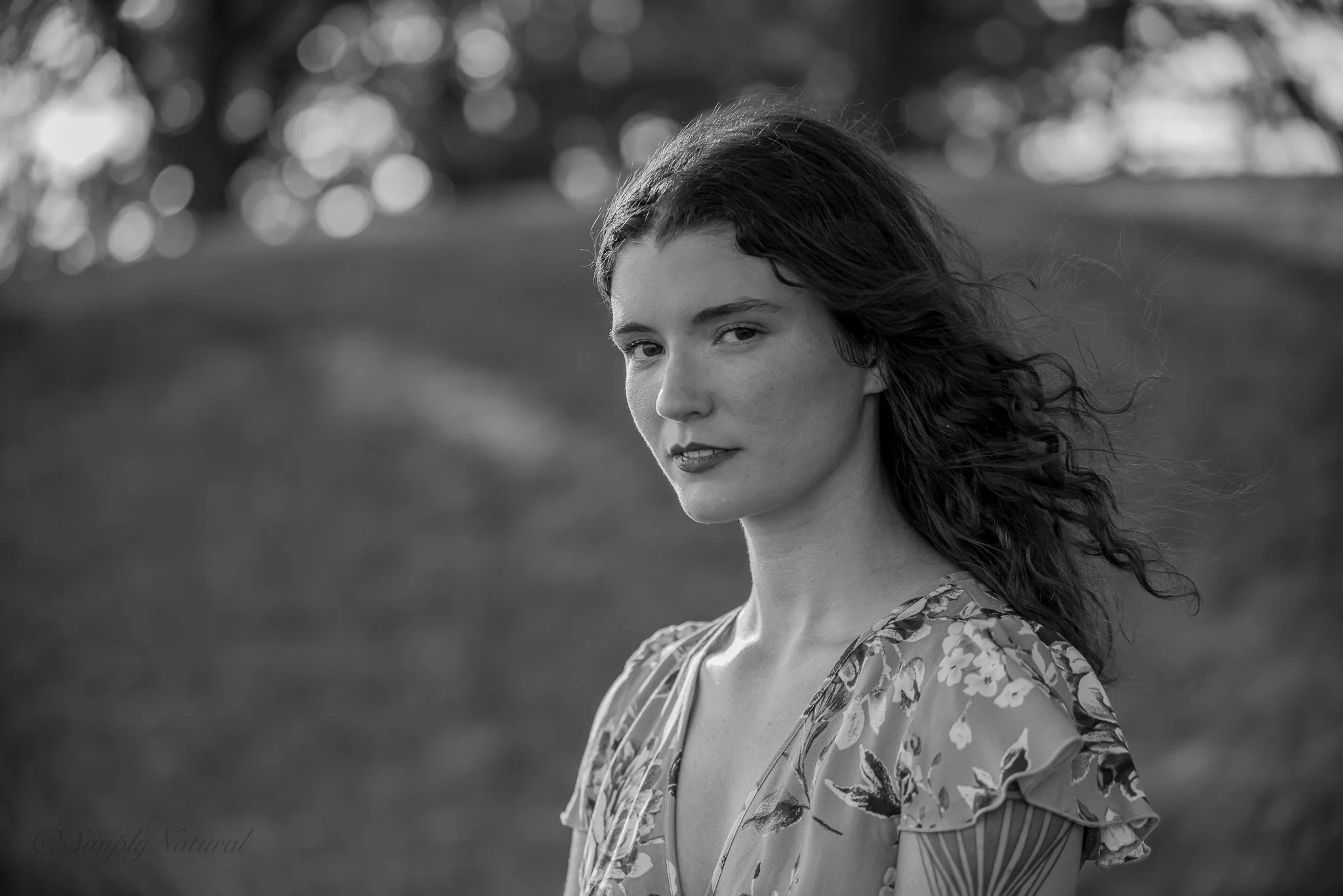 Black and white portrait of a woman with wavy hair, wearing a floral-patterned dress, outdoors in a natural setting.