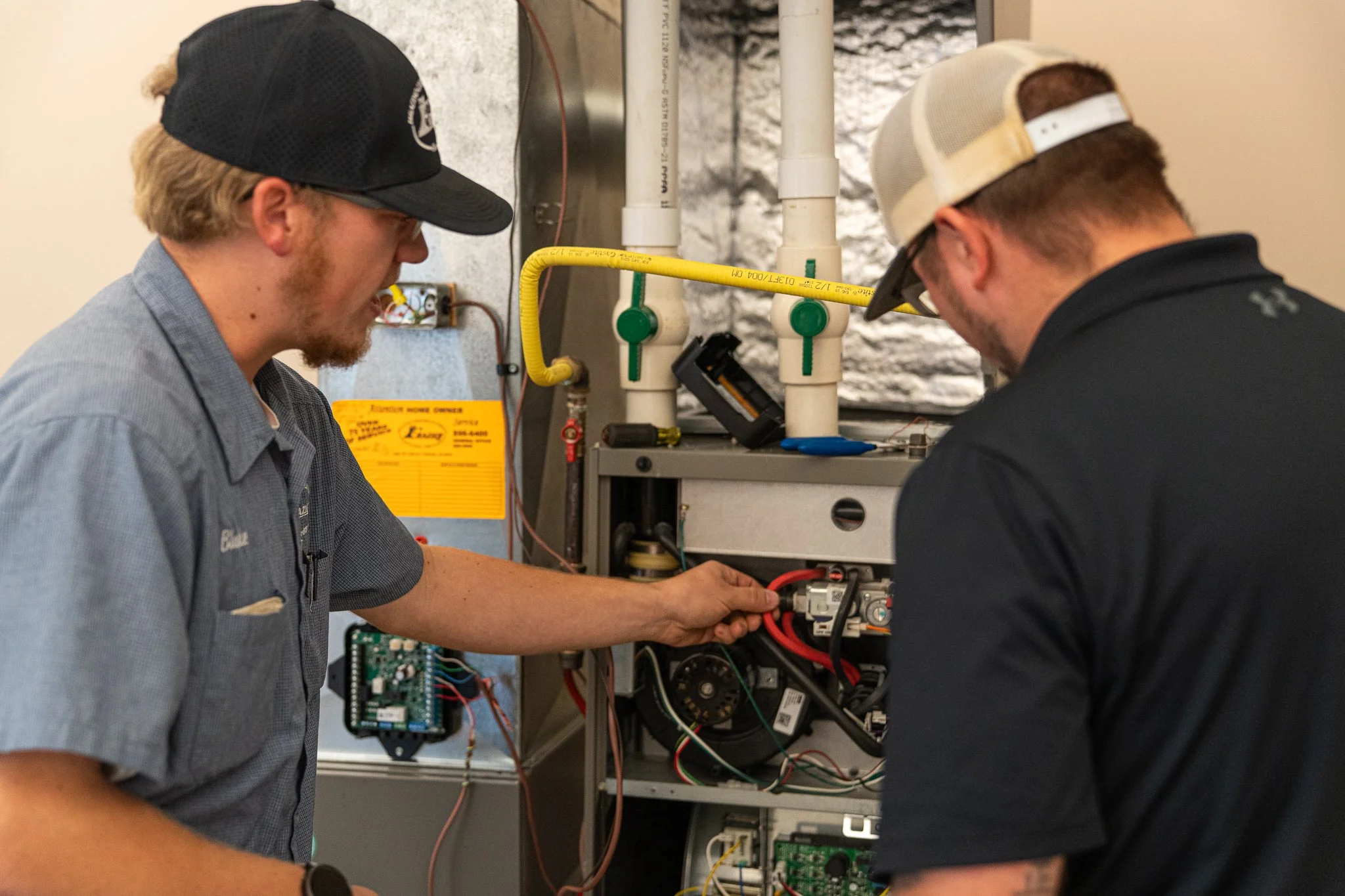Two technicians working on electrical or HVAC equipment, inspecting and repairing components inside an equipment panel, in a professional setting.