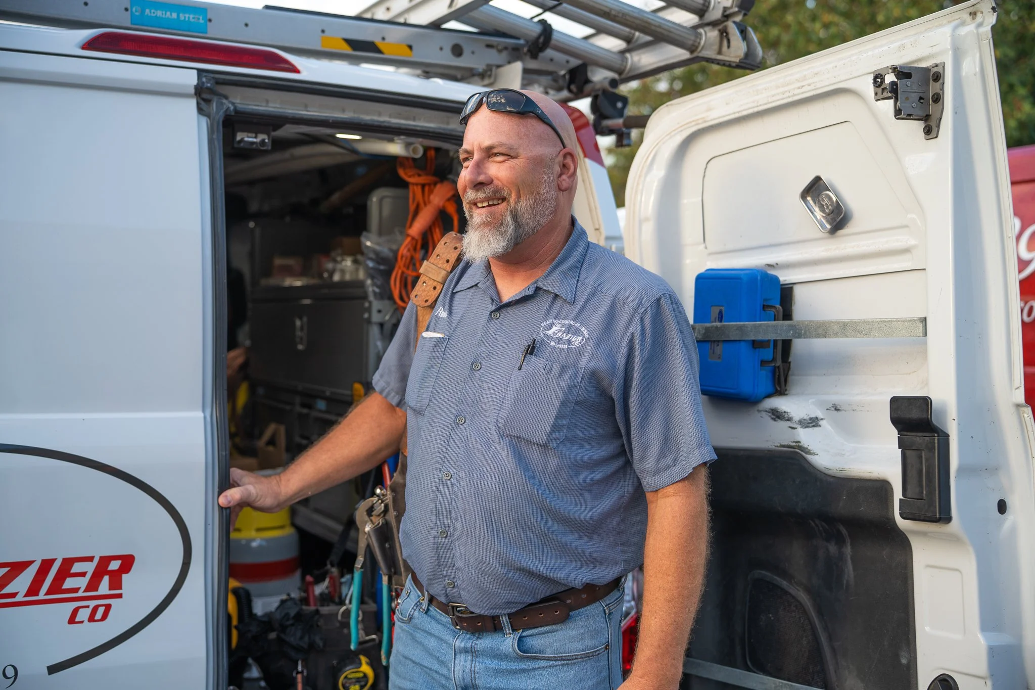 A smiling man with a gray beard and sunglasses on his head stands next to an open work van filled with tools and equipment.