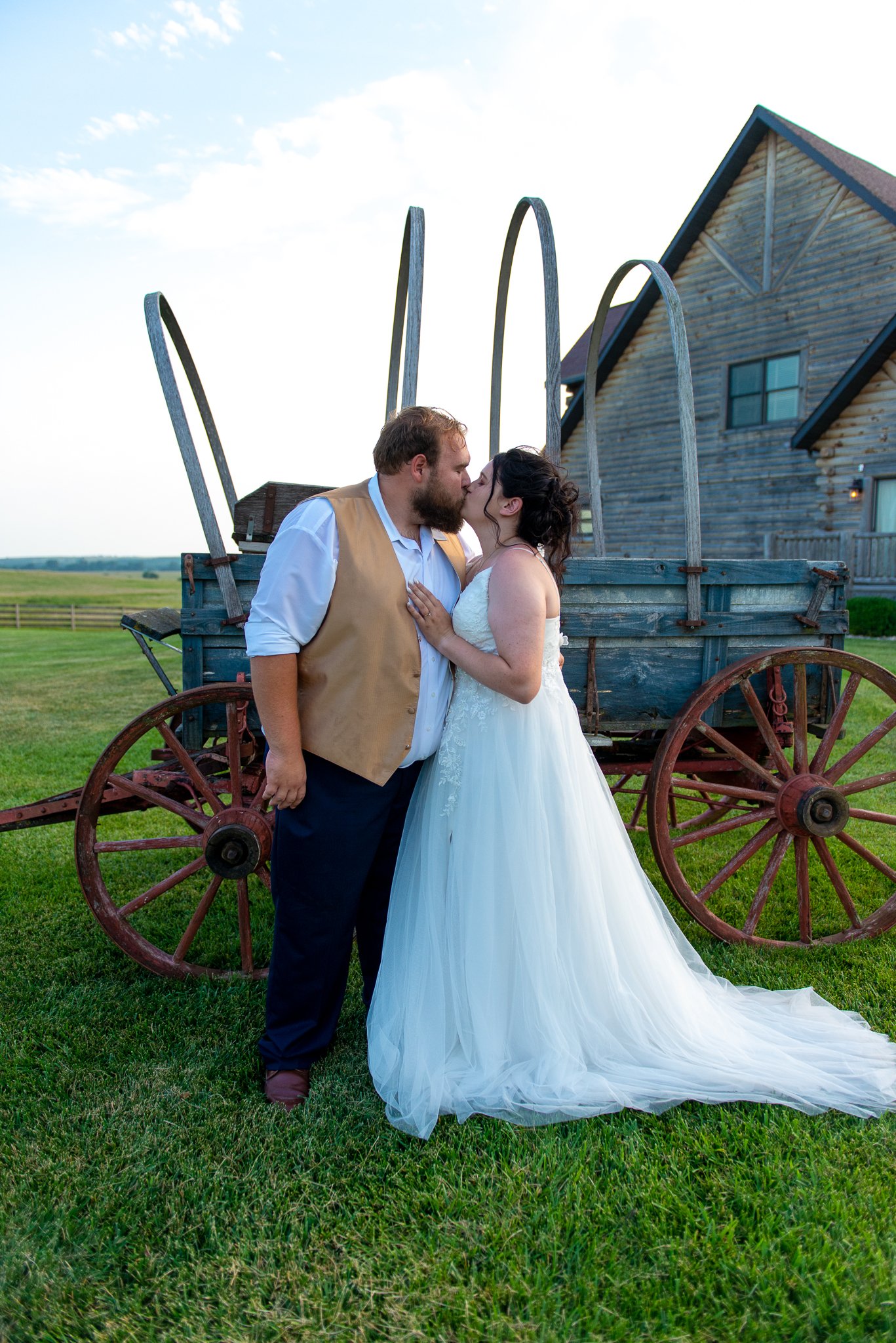A newlywed couple sharing a kiss in front of an old blue wooden wagon and a rustic house, on a grassy field under a partly cloudy sky.