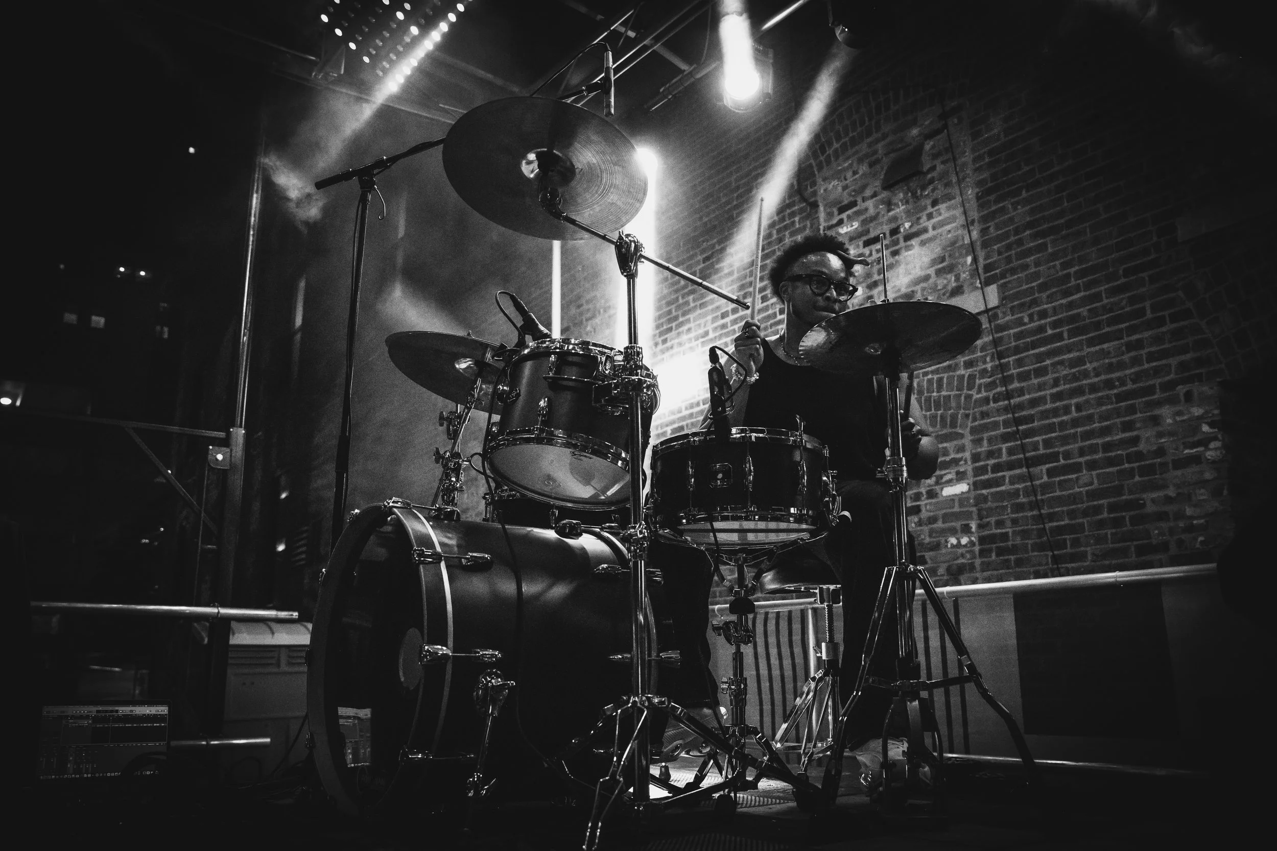 A person playing drums on stage with brick wall background, illuminated by stage lights, in black and white.