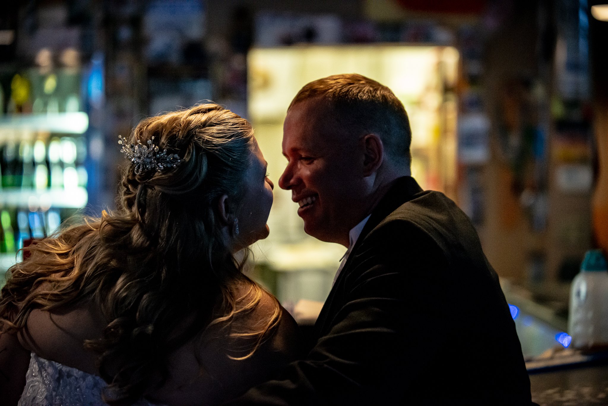 A couple sharing an intimate moment, smiling and leaning close to each other in a dimly lit setting, possibly a bar or restaurant.