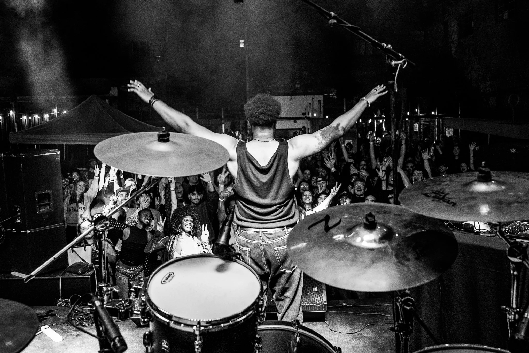 A performer with arms raised on stage during a concert, viewed from behind a drum set, with a crowd of people cheering and raising their hands in the background.