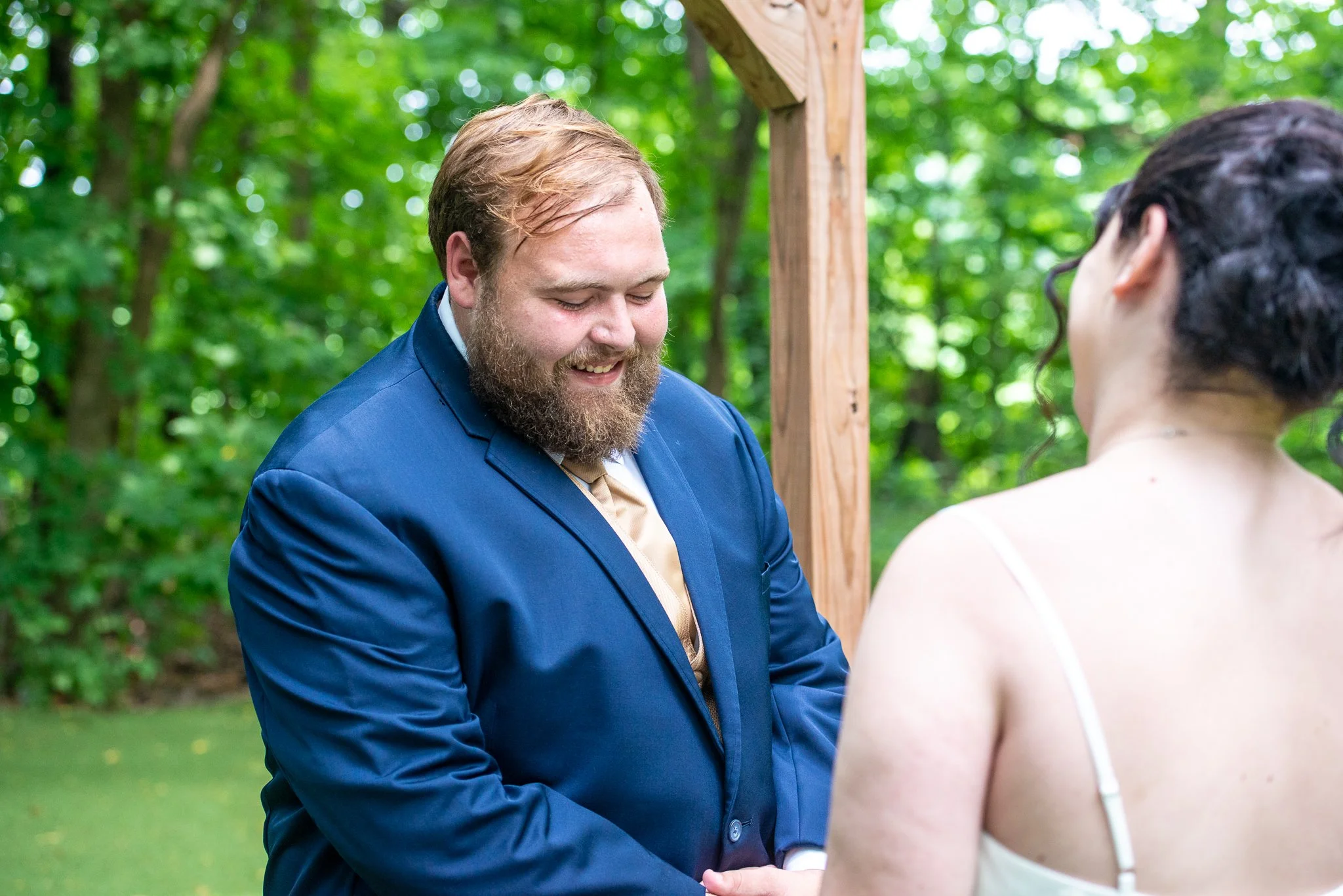 A man in a blue suit appears to be officiating a wedding ceremony with a woman in a white dress outdoors among green trees.