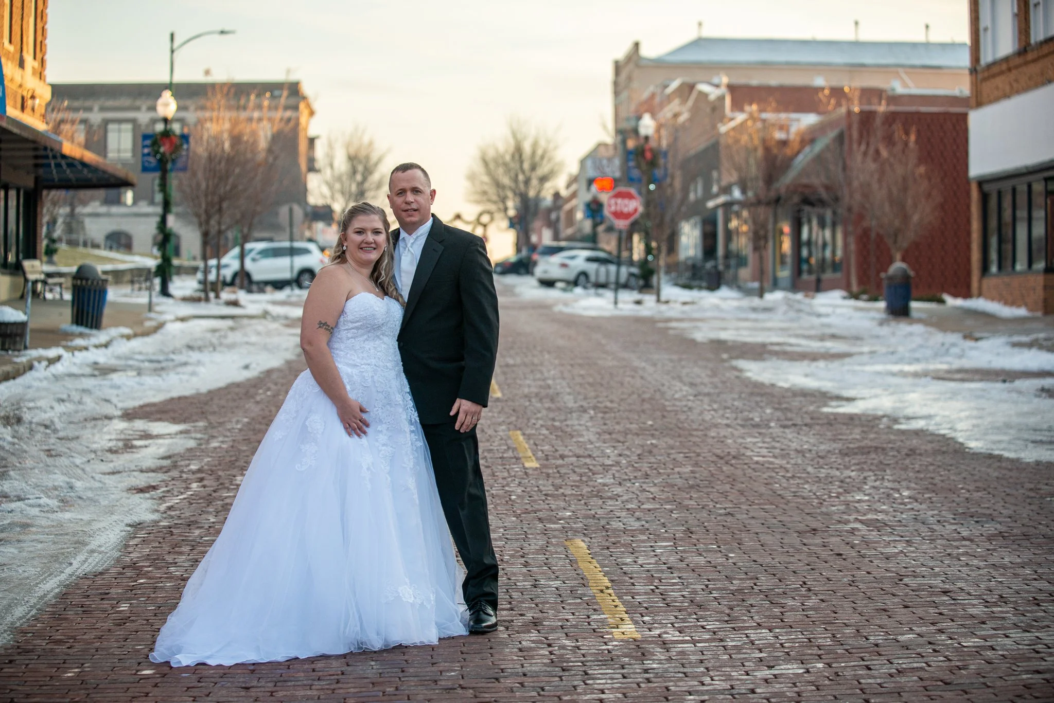 A newlywed couple dressed in wedding attire standing on a cobblestone street in a small town with snow on the ground, brick buildings, and decorated lampposts during sunset.