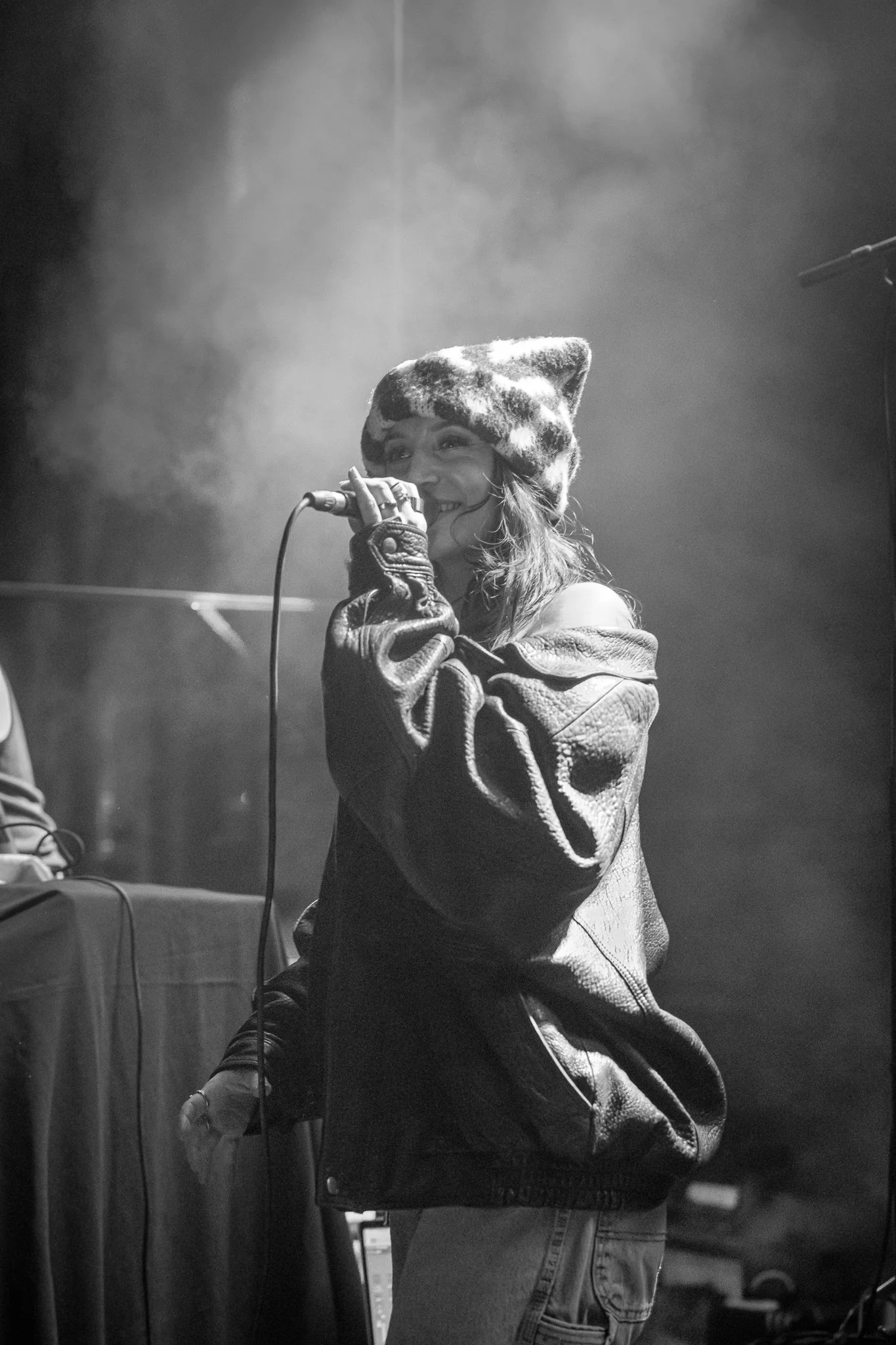A woman with long hair, wearing a hat with a camouflage pattern and a leather jacket, is singing into a microphone in a dimly lit setting.