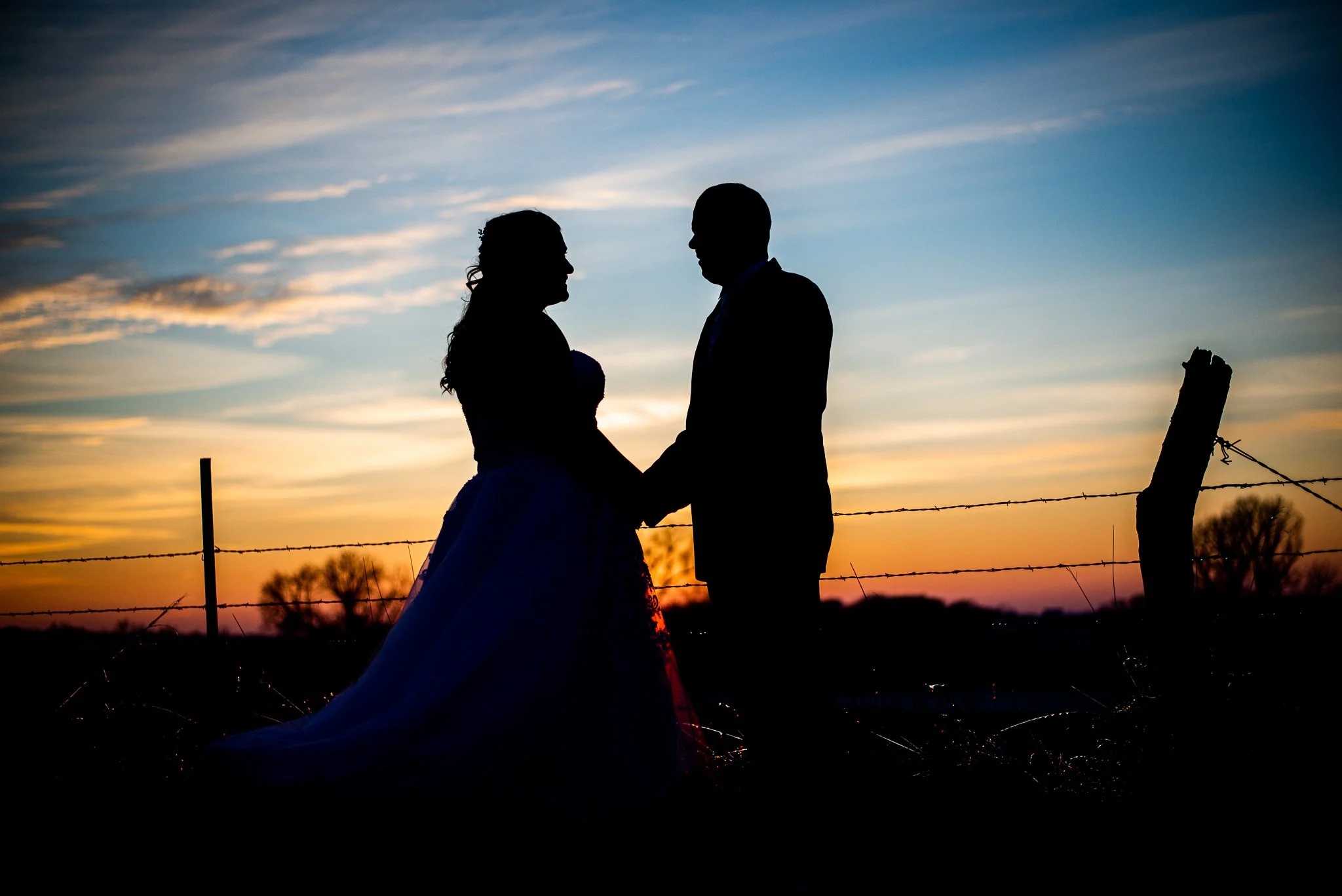 Silhouette of a couple holding hands during sunset, with a sky filled with clouds and a barbed wire fence in the foreground.
