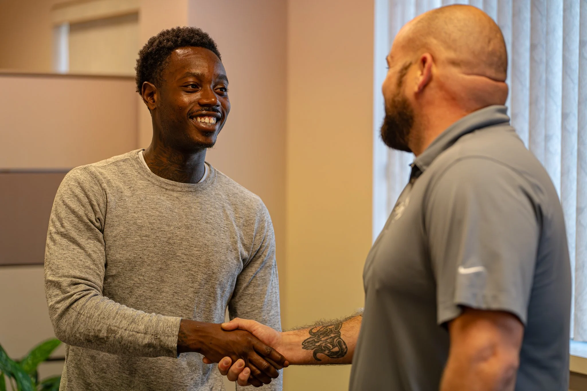 Two men shake hands indoors, one smiling and the other with his back to the camera.