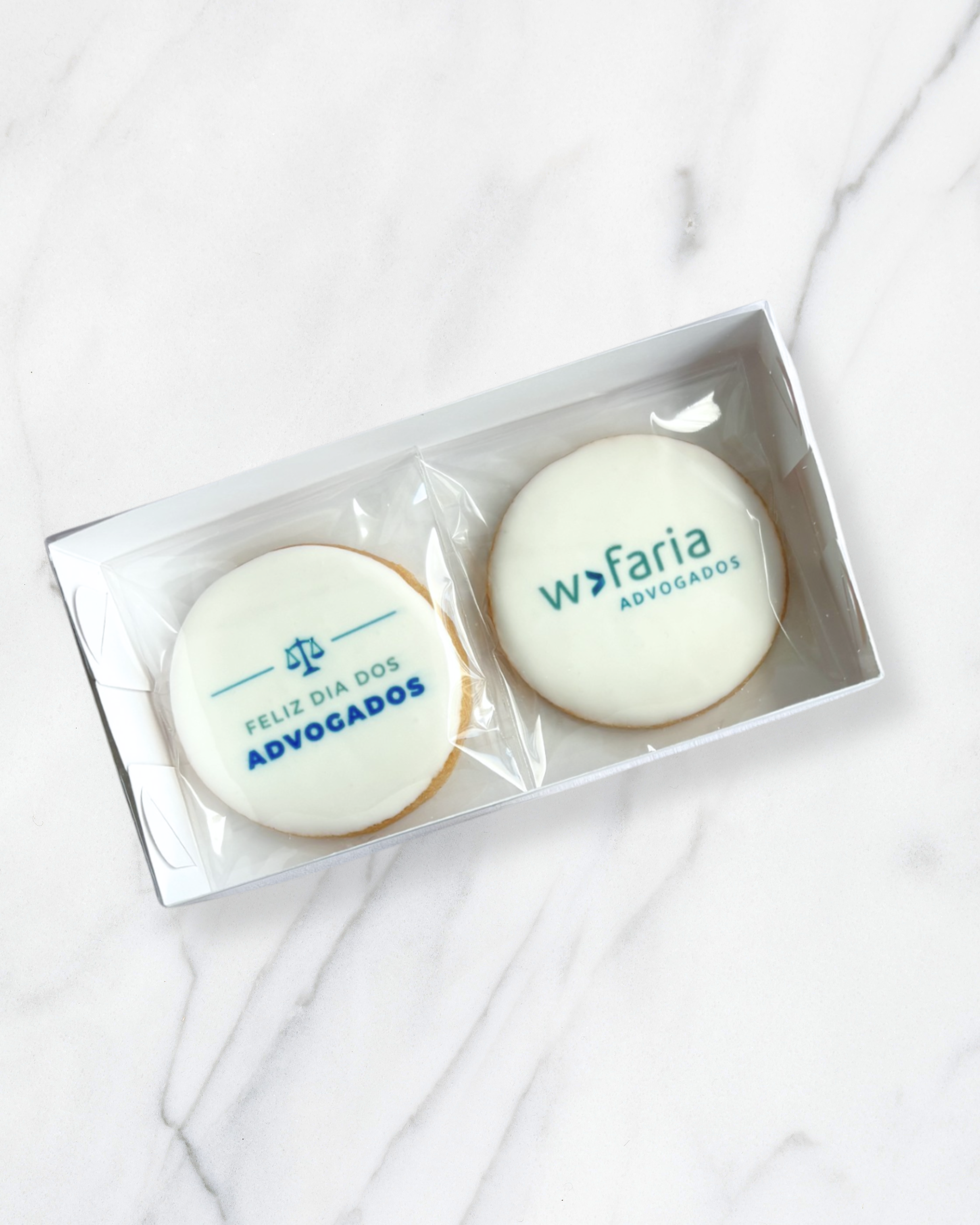 Cookies decorados com mensagem e símbolos em cor azul, embalados individualmente em plasticos transparentes, dentro de uma caixa branca sobre uma superfície de mármore branco. Para uma firma de advocacia.