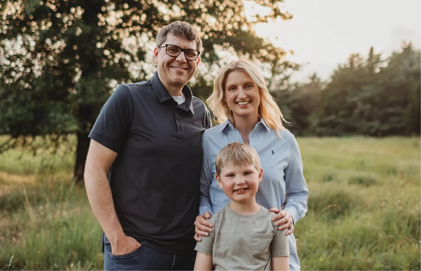 A family of three, including a man, woman, and young boy, standing outdoors in a grassy field with trees in the background, smiling at the camera during sunset.
