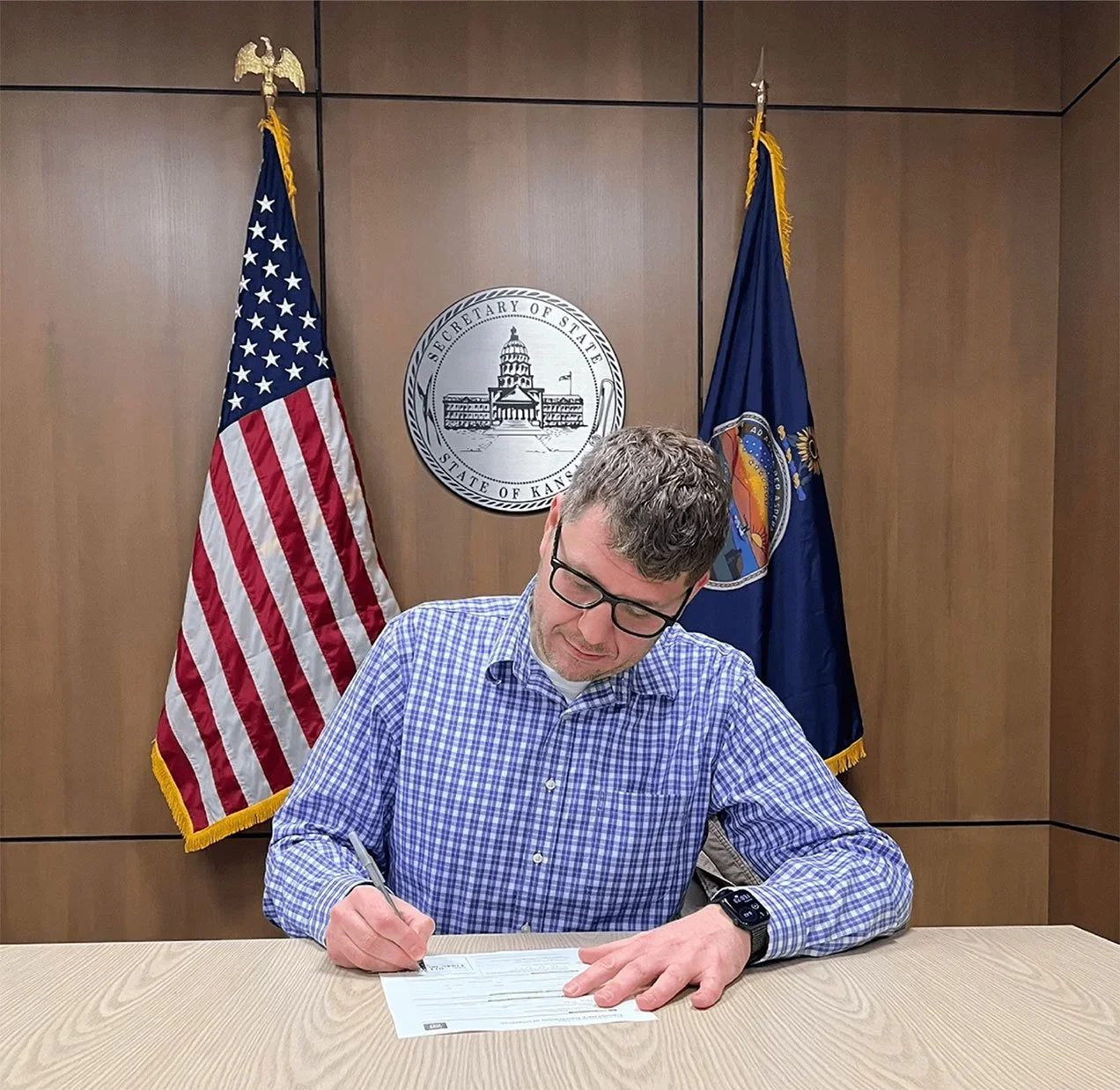 A man with glasses and a checkered shirt signing a document at a table in front of the flags of the United States and Kansas in a government office.