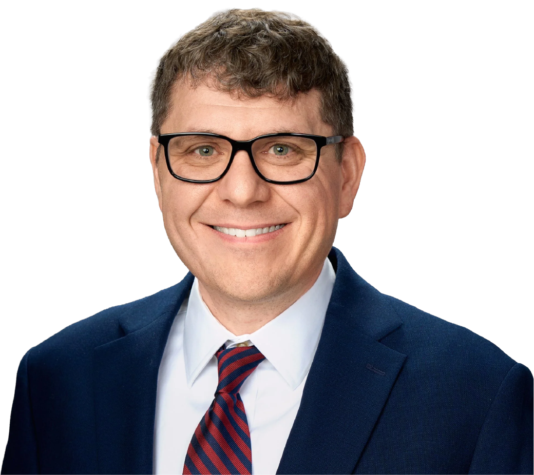 A man in a navy suit, white shirt, and striped red and blue tie, smiling and wearing black glasses.