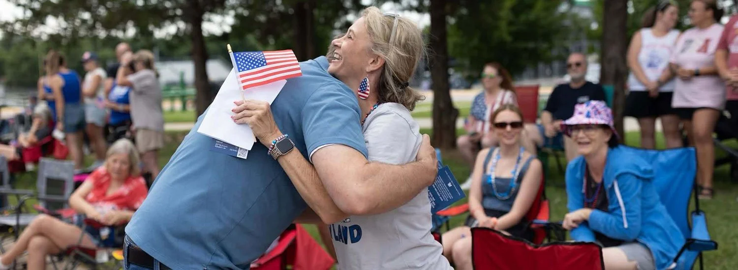 Two women embrace at an outdoor gathering, with one woman wearing a blue shirt holding American flags, and several people sitting and standing in the background in a park setting.
