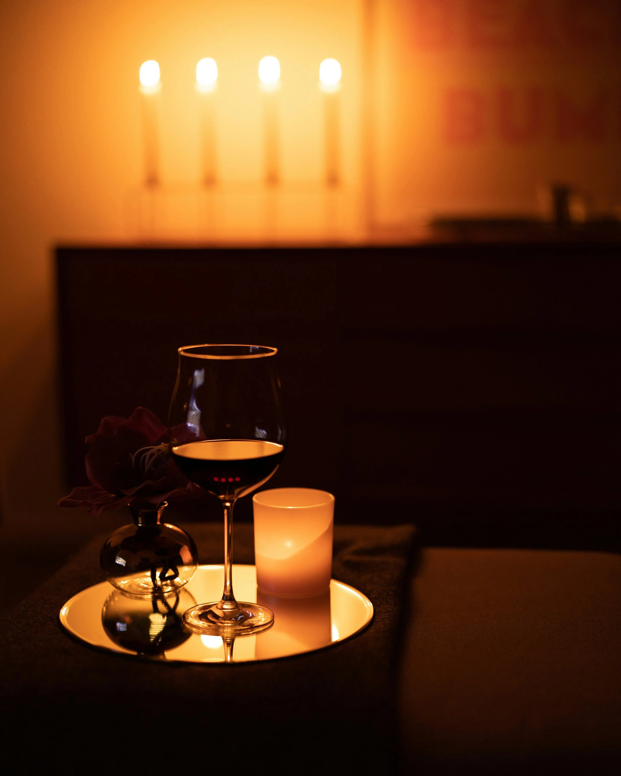 A glass of red wine, a small candle, and a vase with dark flowers on a tray, illuminated by warm candlelight, with a candelabra in the background.