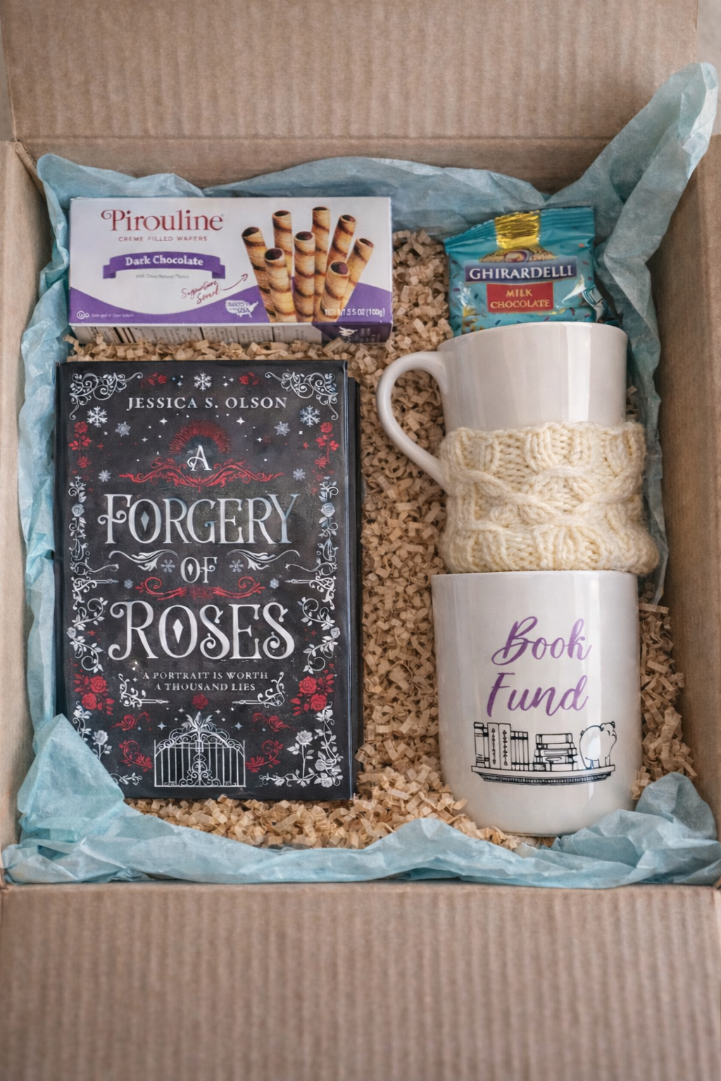 A gift box with a book titled 'A Forgery of Roses', a white mug with a knitted cozy, another mug with 'Book Fund' written on it, a box of dark chocolate Pirouline wafers, a Ghirardelli milk chocolate bar, and a white ceramic cup with a handle, all ar