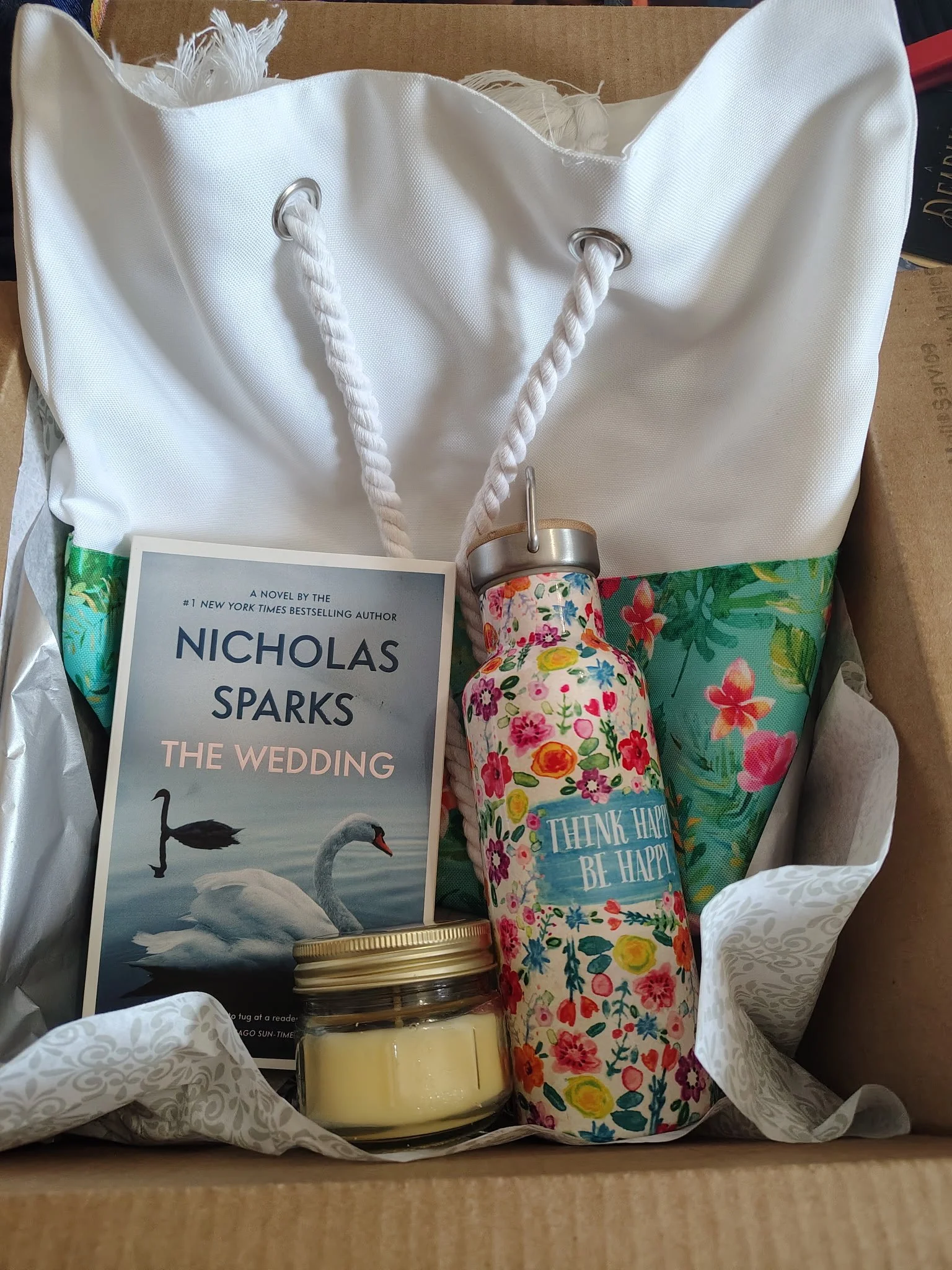 A gift box containing a novel titled "The Wedding" by Nicholas Sparks, a floral water bottle with a "Think Happy Be Happy" label, a jar of yellow candle, and a colorful fabric with a floral pattern.
