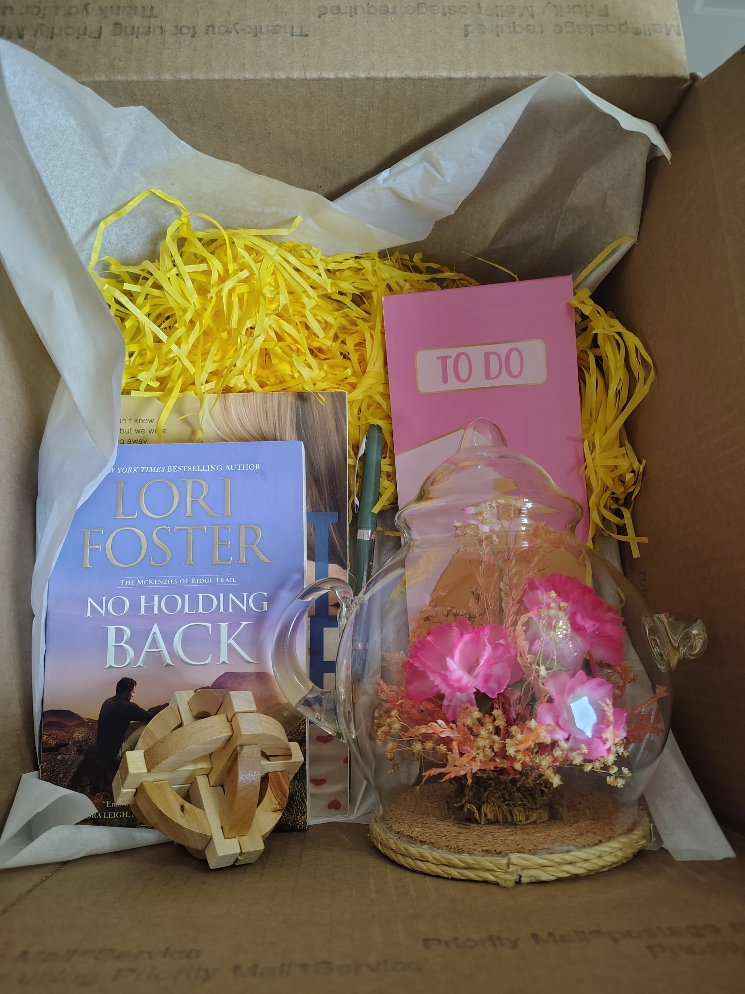 A gift box containing a paperback book titled 'No Holding Back' by Lori Foster, a pink 'TO DO' notepad, a glass cloche with pink flowers inside, a wooden geometric heart, a green pen, and yellow shredded paper lining the box.