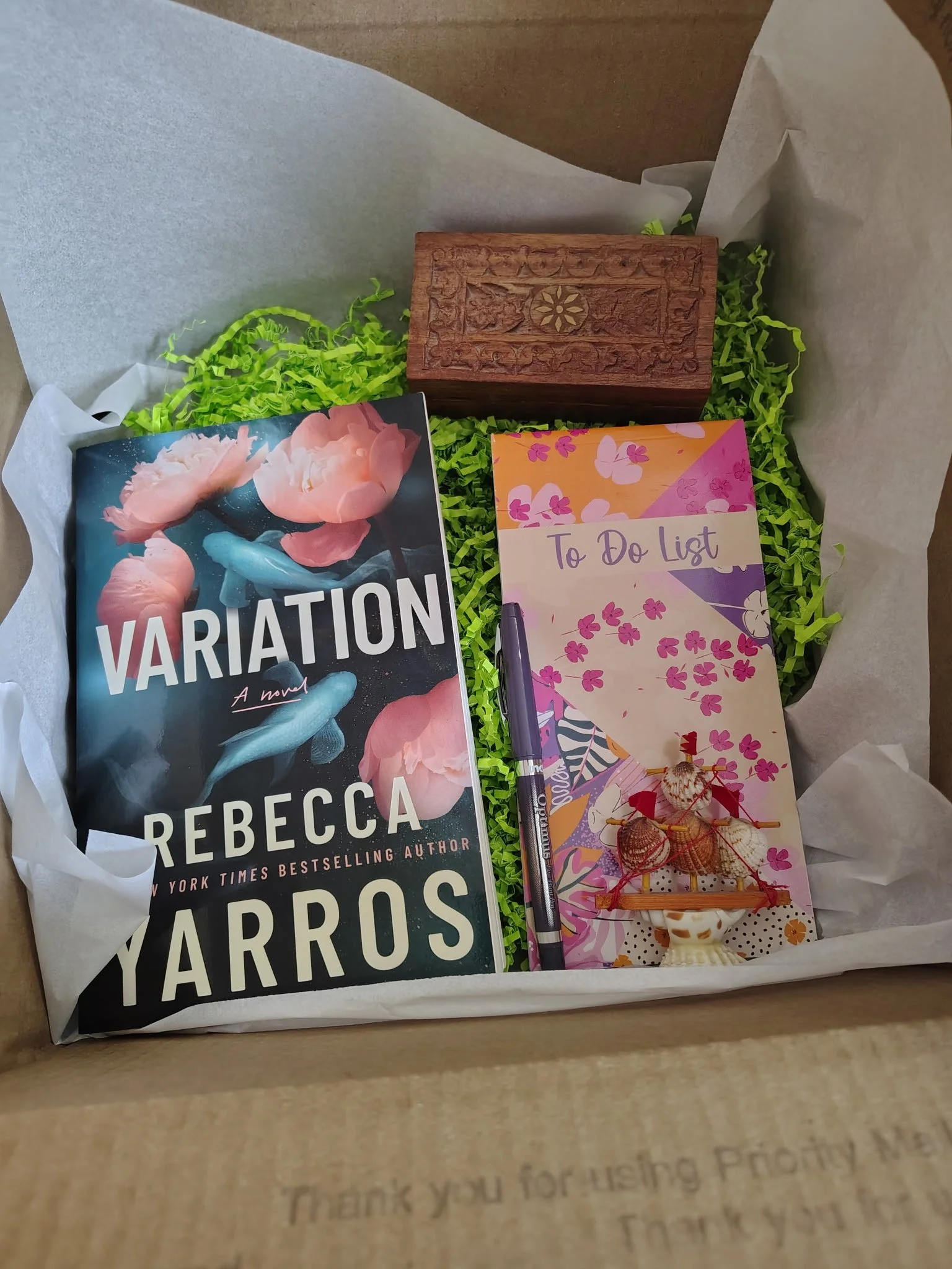 Open box containing a copy of the book "Variation" by Rebecca Yarros, a to-do list notepad, a decorative wooden box, a purple pen, and a small ship model.