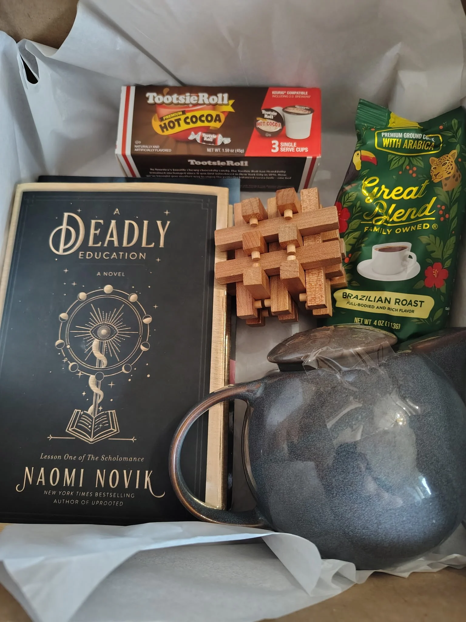 A box containing a book titled 'A Deadly Education' by Naomi Novik, a package of Ground Coffee, a box of Tootsie Rolls, a ceramic pitcher, and a wooden puzzle.