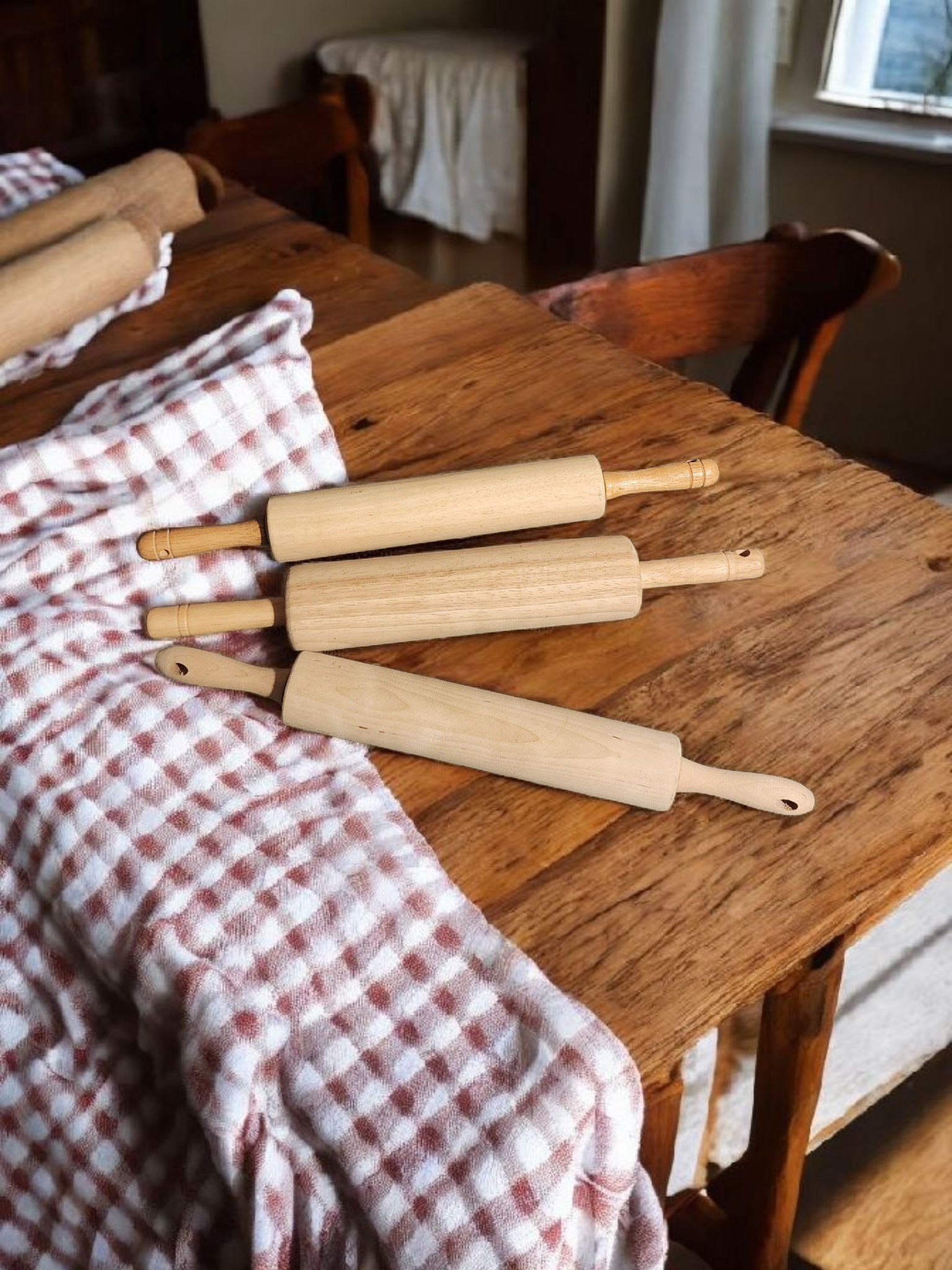 Three wooden rolling pins placed on a rustic wooden table covered with a red and white checkered cloth in a cozy kitchen setting.