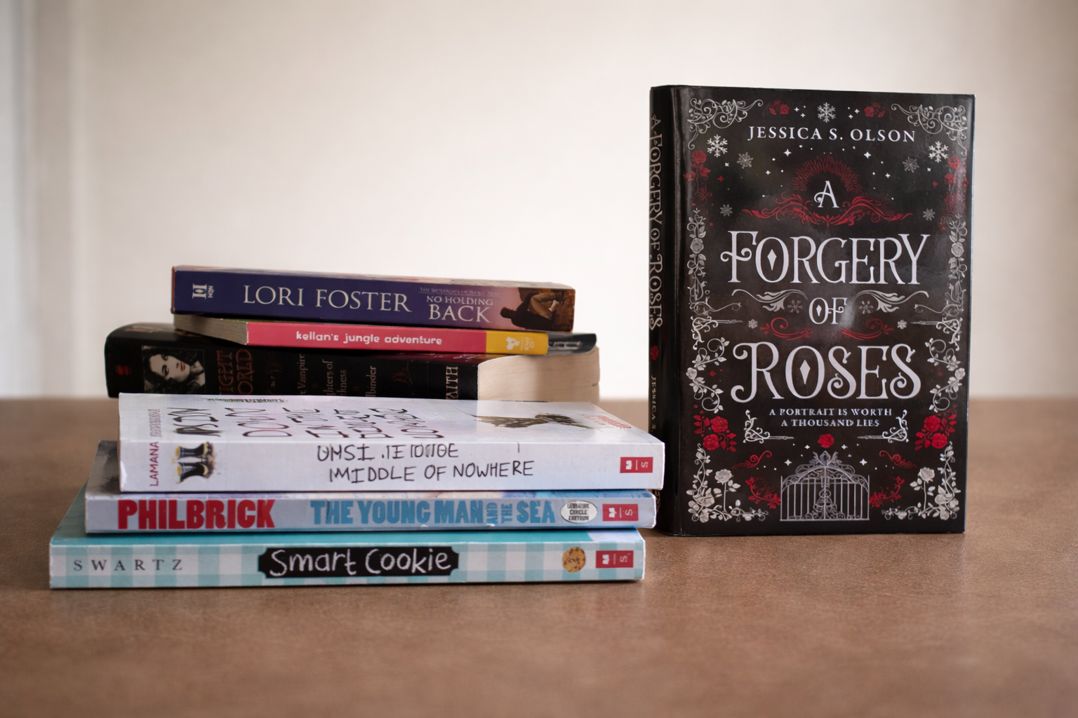 A stack of seven books on a wooden table with a black and red book titled 'A Forgery of Roses' standing upright beside the pile.