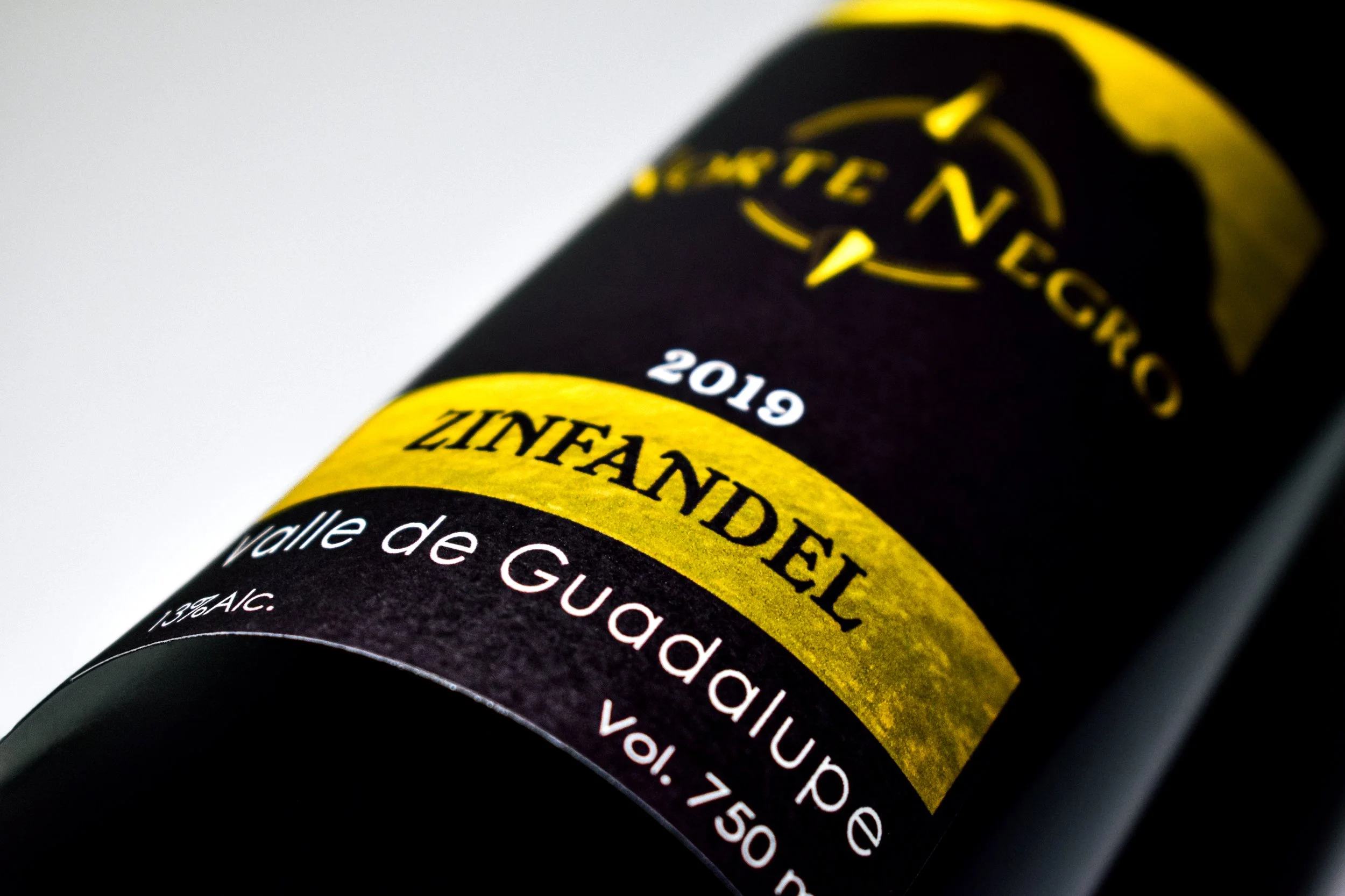 Botella de vino tinto Zinfandel con etiqueta negra, amarilla y blanca, que indica Valle de Guadalupe, 2019, volumen de 750 ml.
