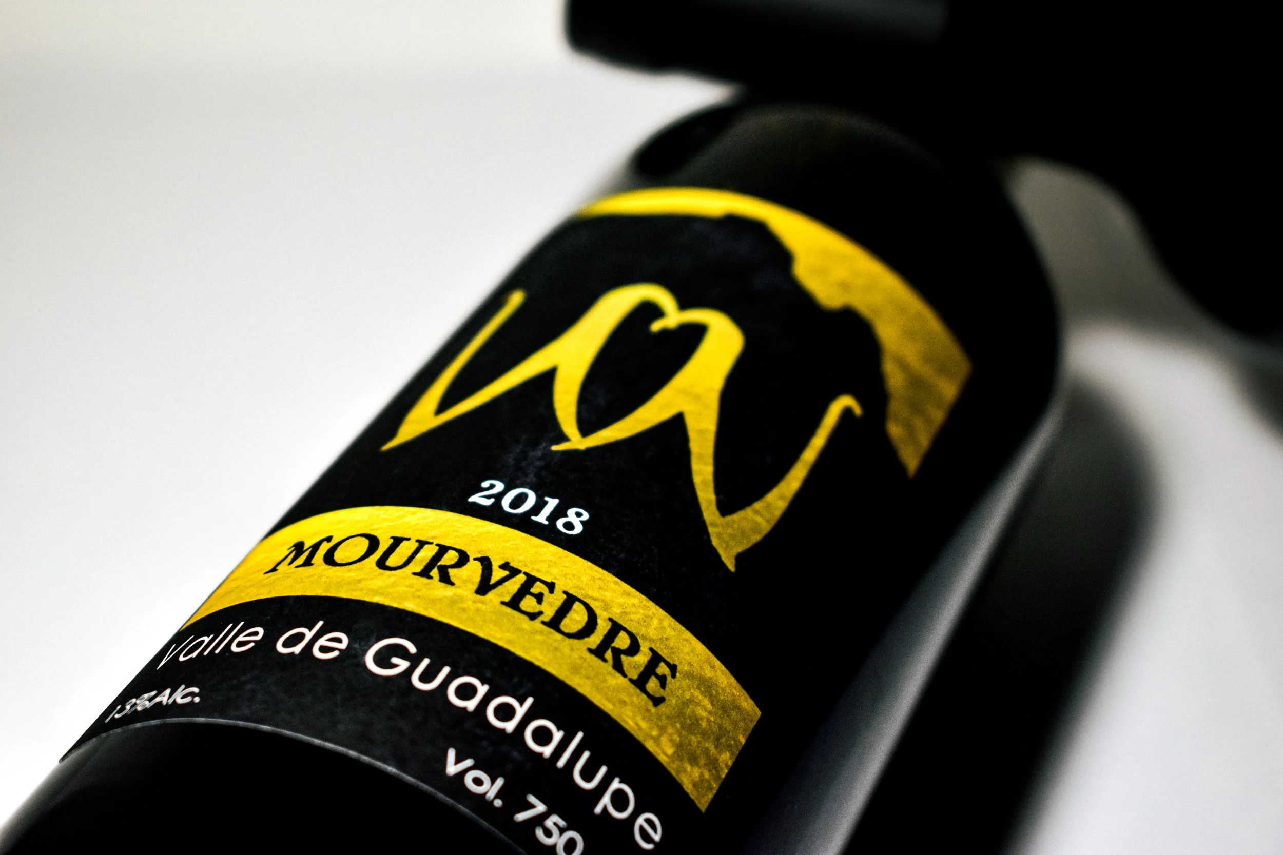 Botella de vino tinto con etiqueta negra y dorada que dice 'Vivu 2018' y 'Mourvèdre' de Valle de Guadalupe.