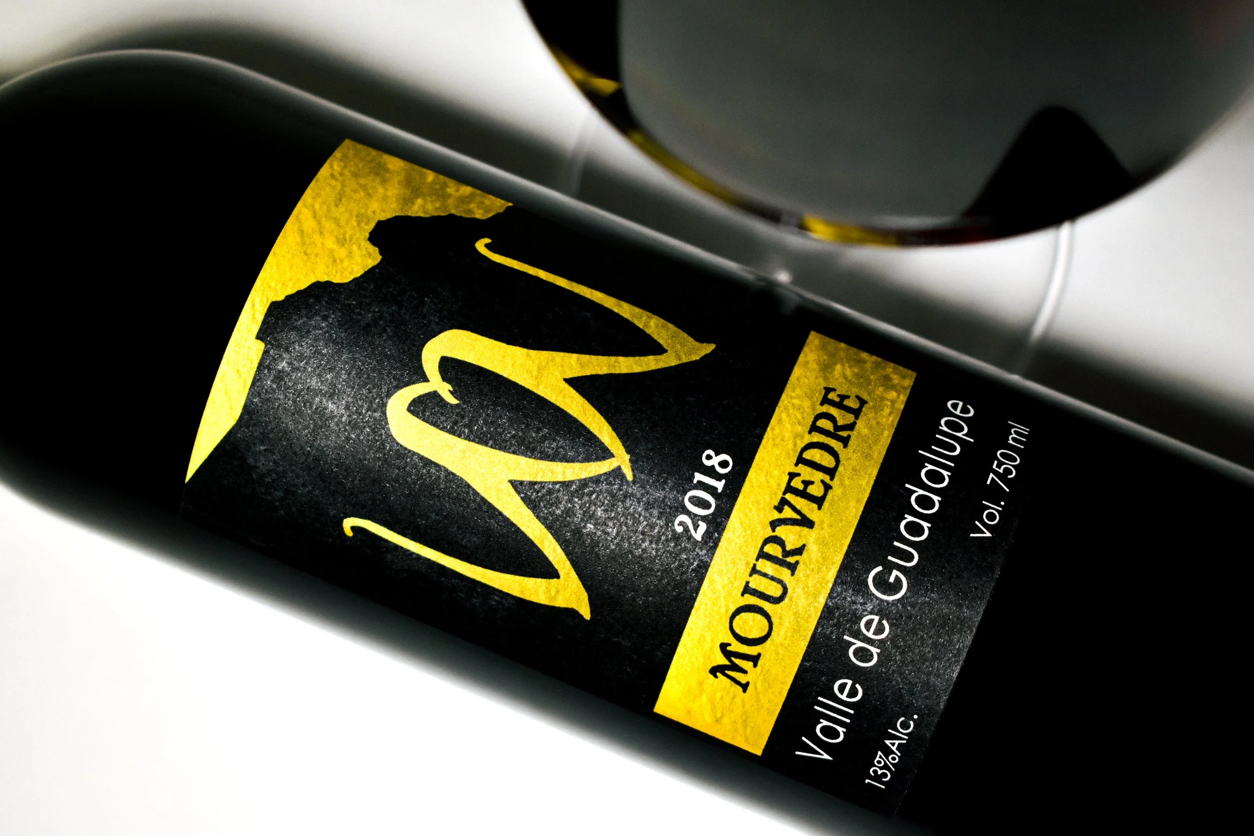 Botella de vino tinto con etiqueta negra y amarilla, marca Mourvèdre, de la cosecha 2018, volumen de 750 ml, con 13% de alcohol.