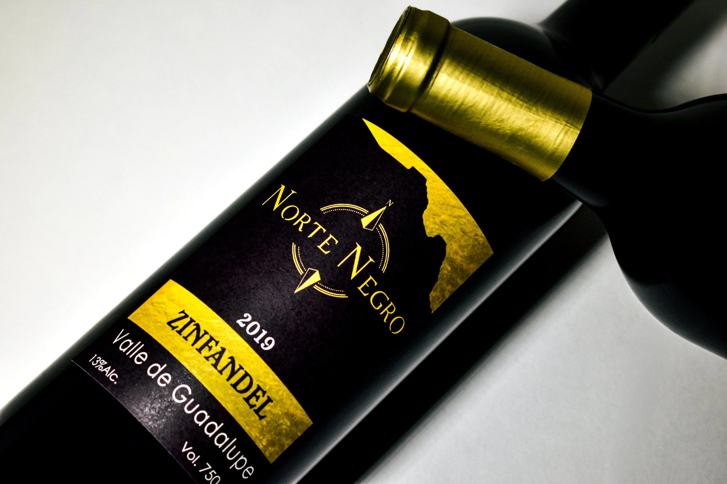 Botella de vino tinto con etiqueta negra, dorada y amarilla en un fondo blanco. La etiqueta indica que es de la región Valle de Guadalupe, producción del 2019, de la variedad Zinfandel, con 13% de alcohol.