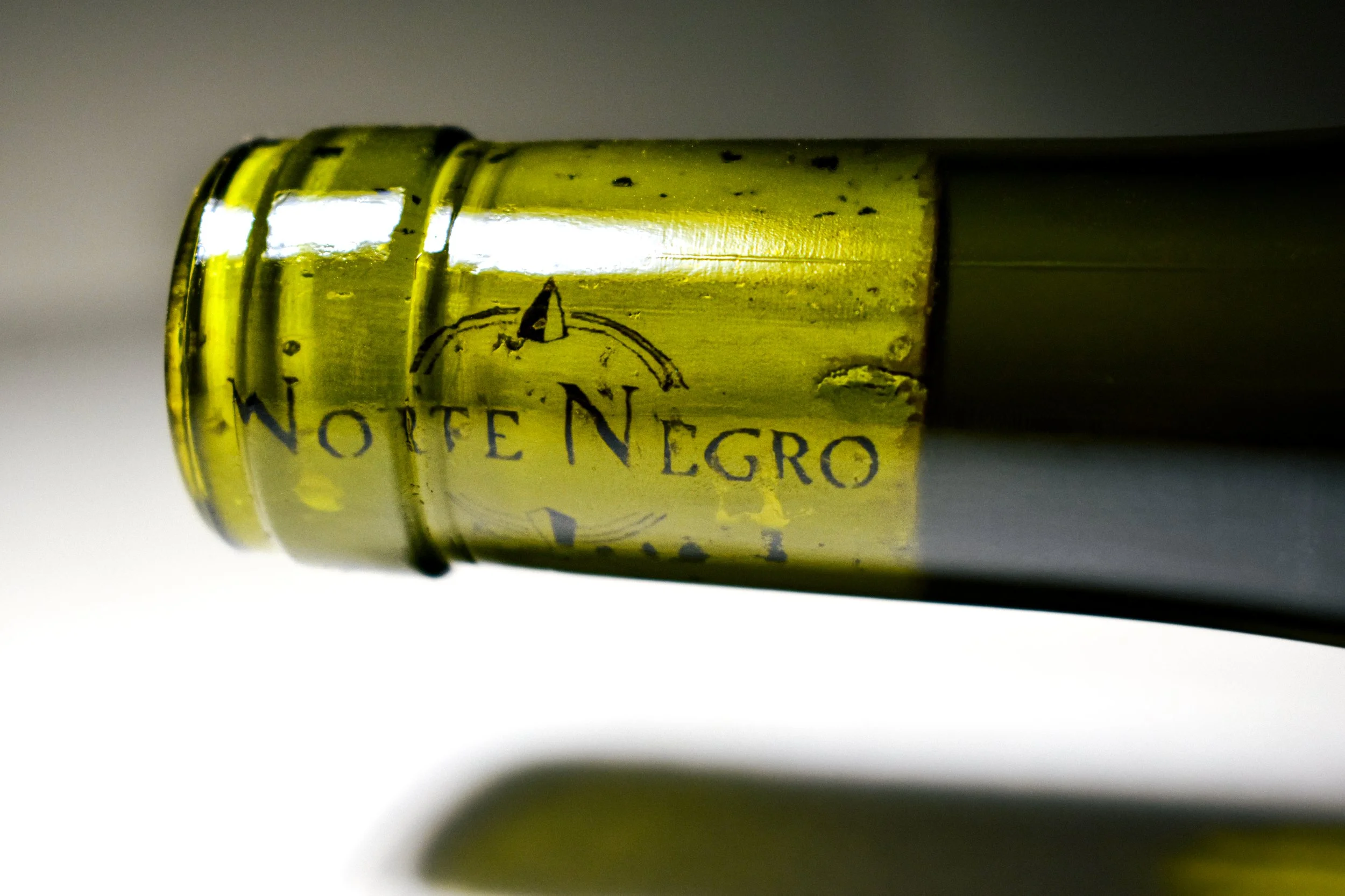 Botella de vino blanca con etiqueta que dice 'Note Negro' en letras negras, vista lateral desde abajo, con reflejos y fondo desenfocado.