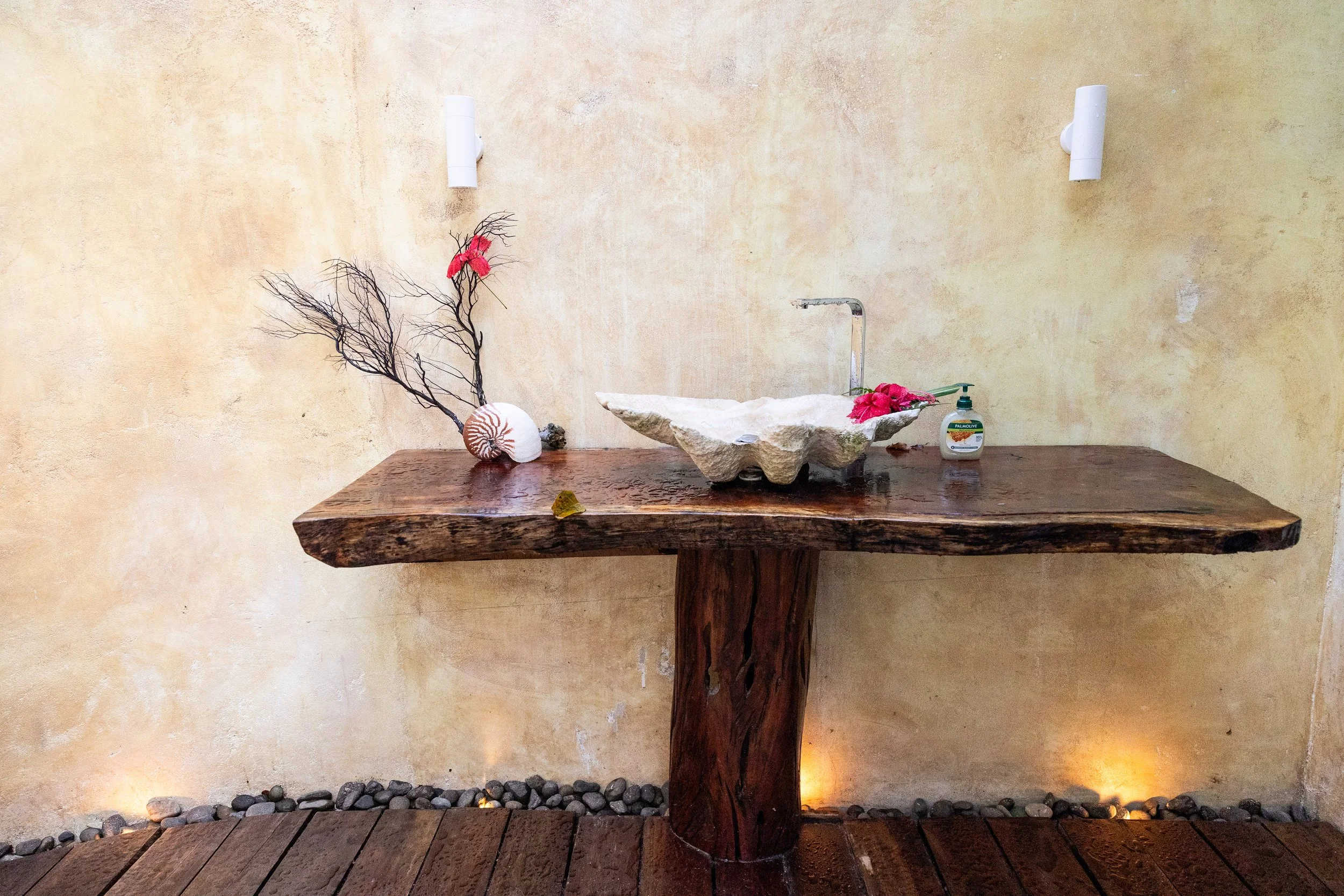 Rustic wooden sink with a large stone basin, decorated with pink flowers, and a soap dispenser on the right, against a beige textured wall with two wall sconces and a wooden floor with small rocks at the base.