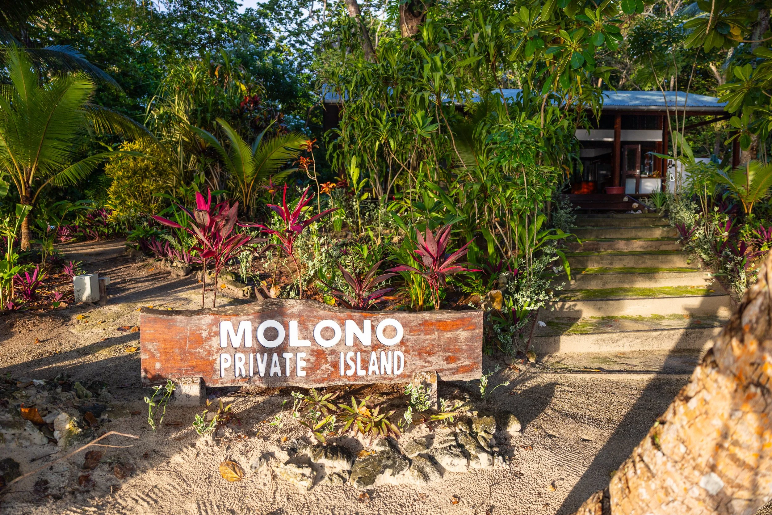 Wooden sign reading 'Molono Private Island' in front of tropical plants and steps leading to a small structure surrounded by lush greenery.