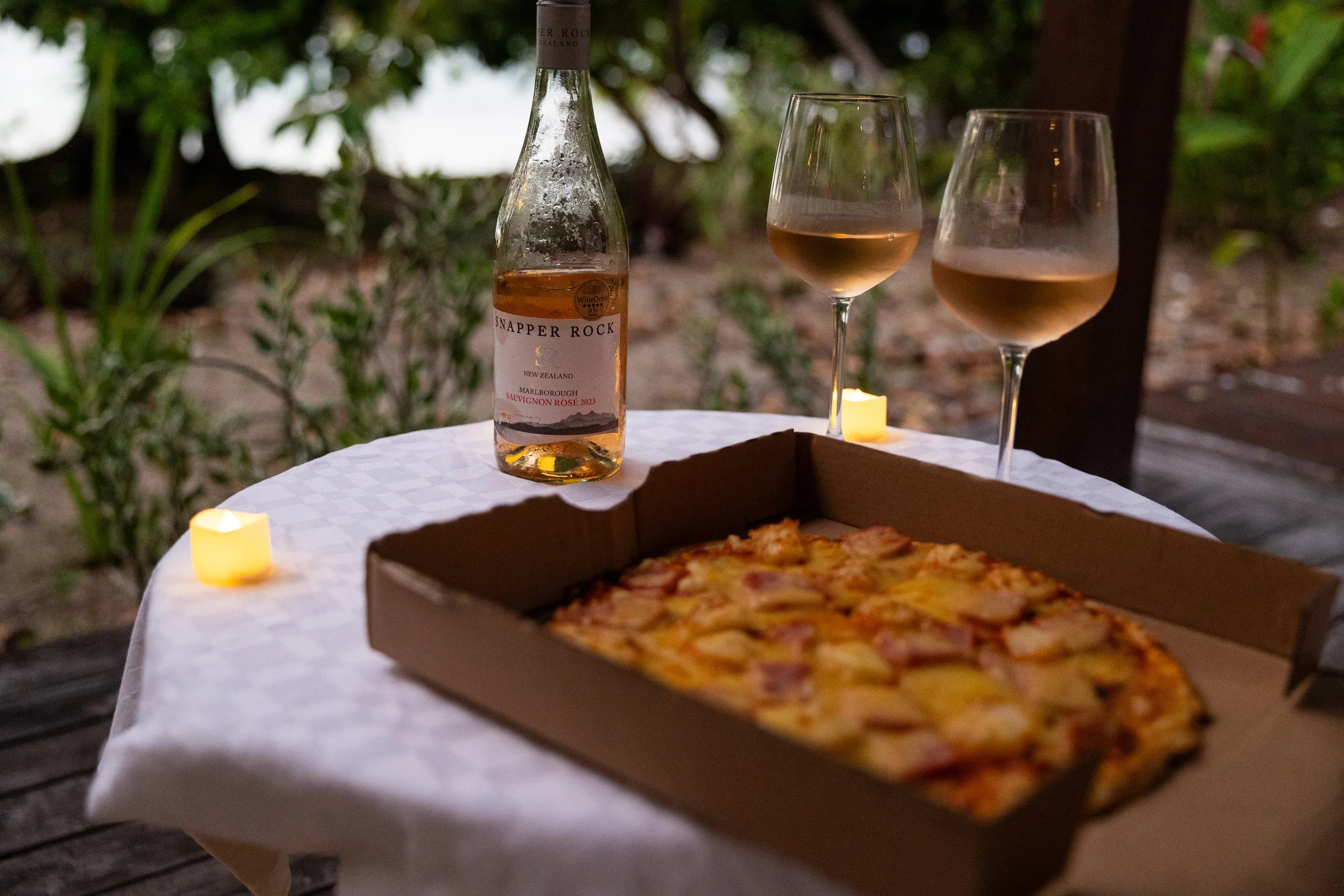 A pizza in a cardboard box, a bottle of wine, and two glasses of rosé wine on a table with a white tablecloth, lit by small candles outdoors in a garden at dusk.