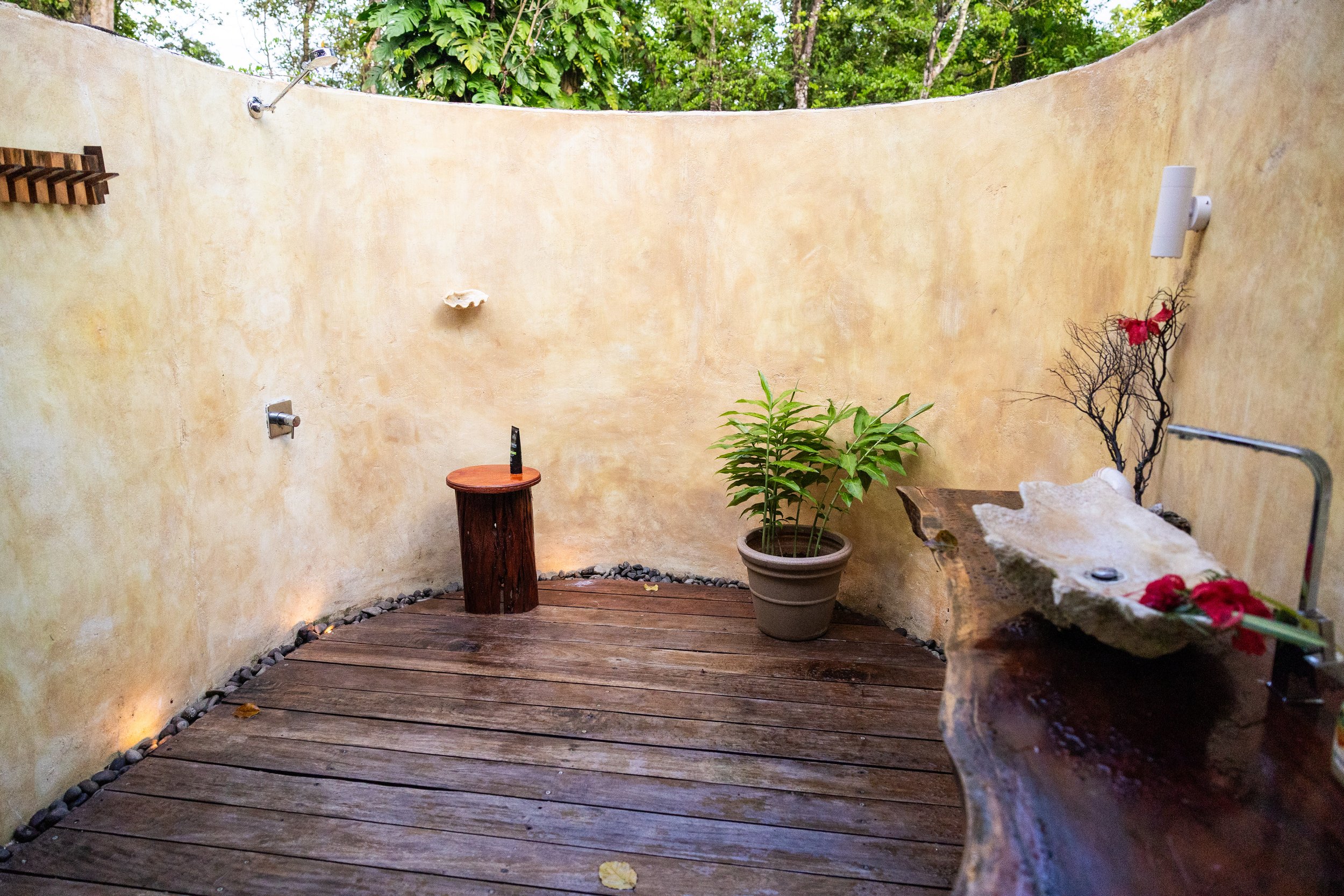 Outdoor shower area with a curved sandstone wall, wooden floor, potted plant, and a clamshell sink with red flowers, surrounded by trees.